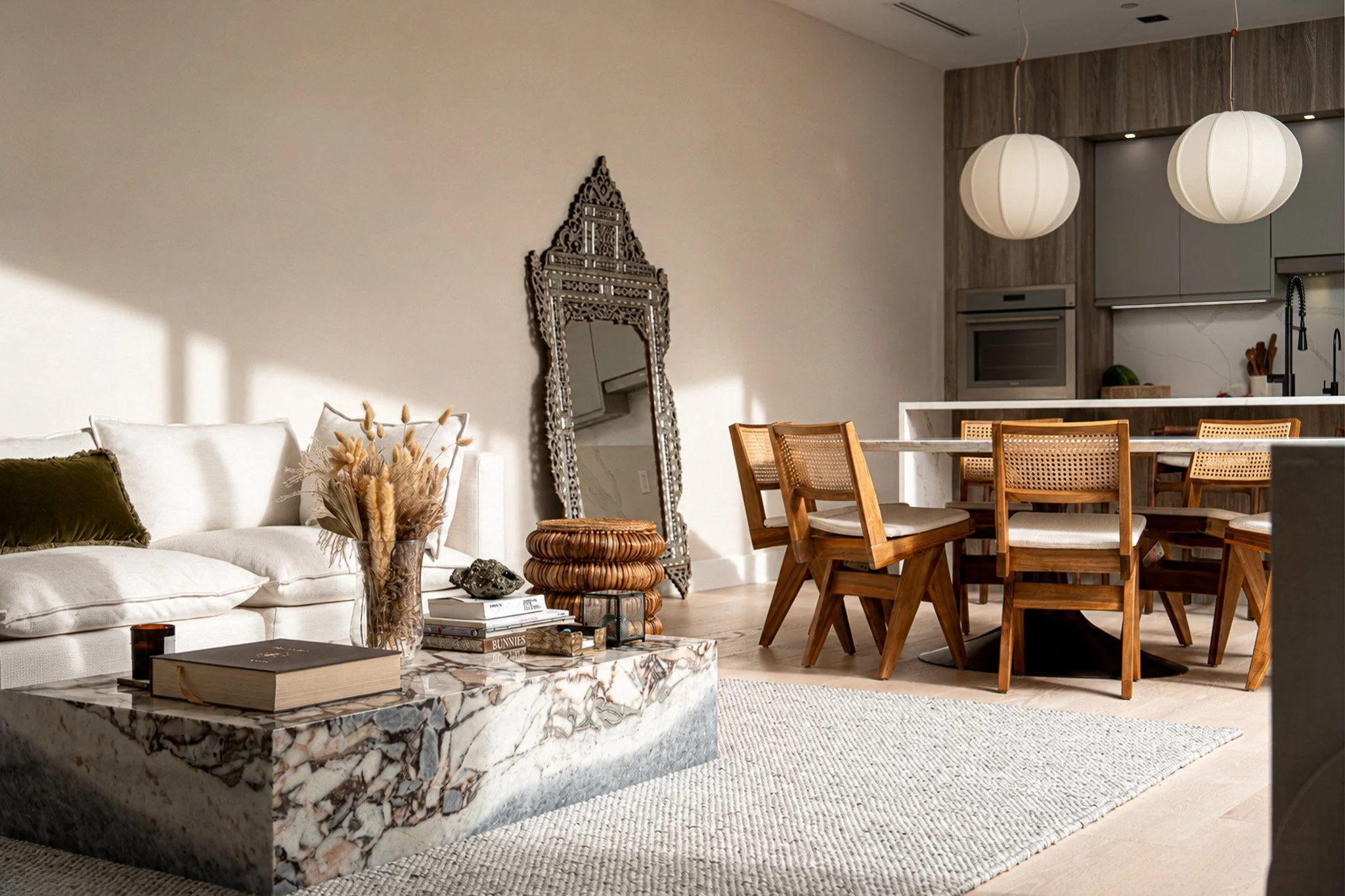 Modern living room and dining area with a white sofa, decorative mirror, marble coffee table, wooden dining chairs, pendant lights, and kitchen in the background.