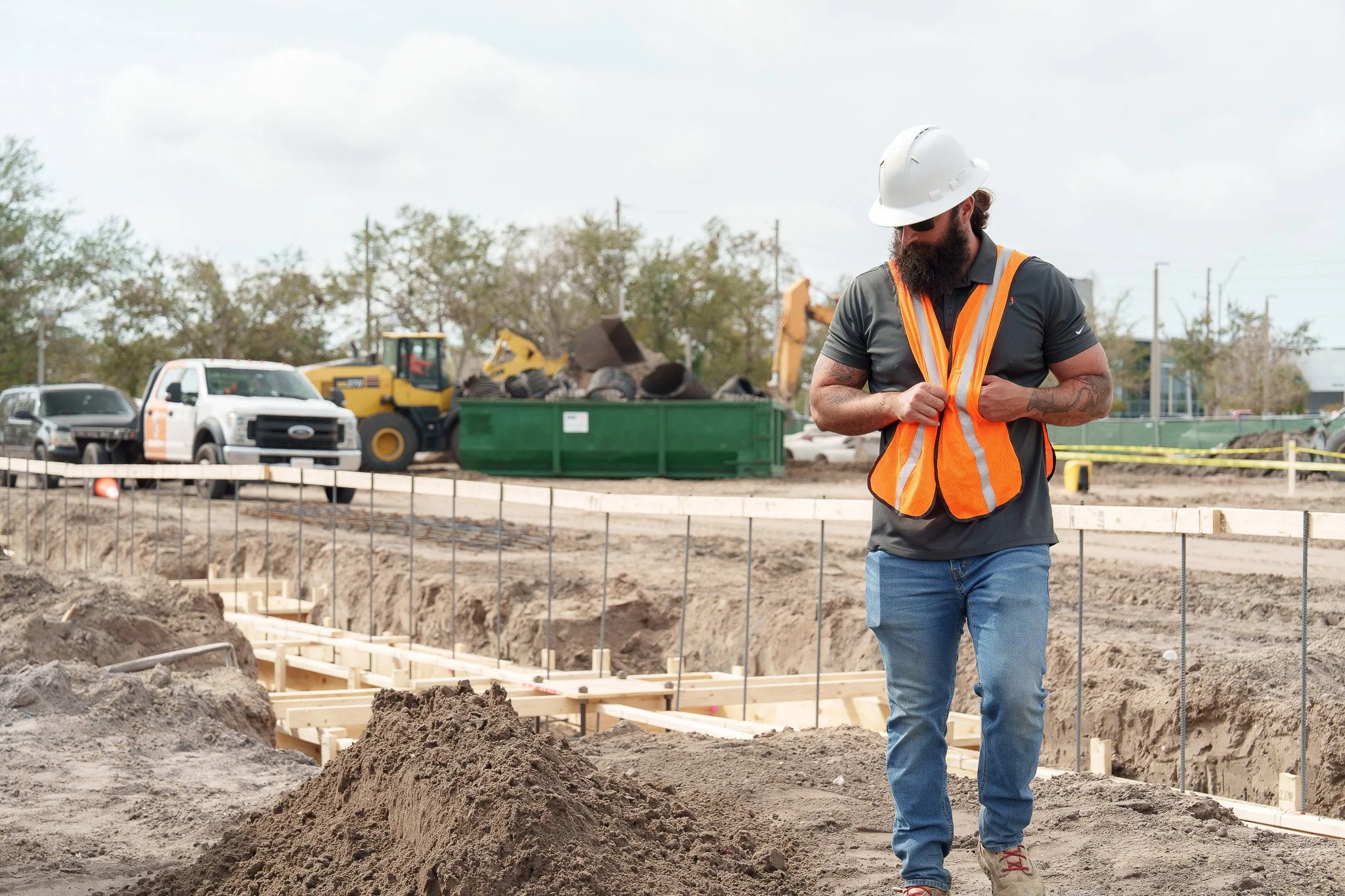A construction site with a man wearing a white hard hat, sunglasses, an orange safety vest, and jeans, standing among dirt and wooden framing. In the background, there are construction vehicles and trees.