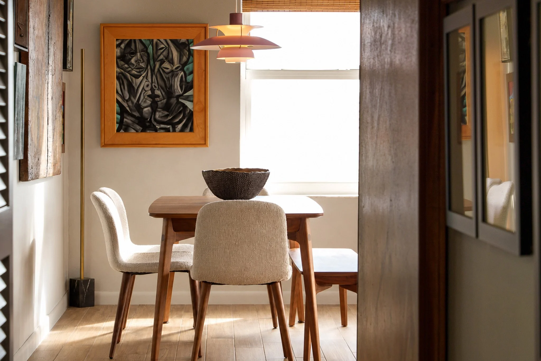 A cozy dining room with a wooden table and beige upholstered chairs. A large black woven bowl is on the table. Natural light comes through a window with a wooden blind. A colorful abstract painting hangs on the wall, and a modern pink and white chand