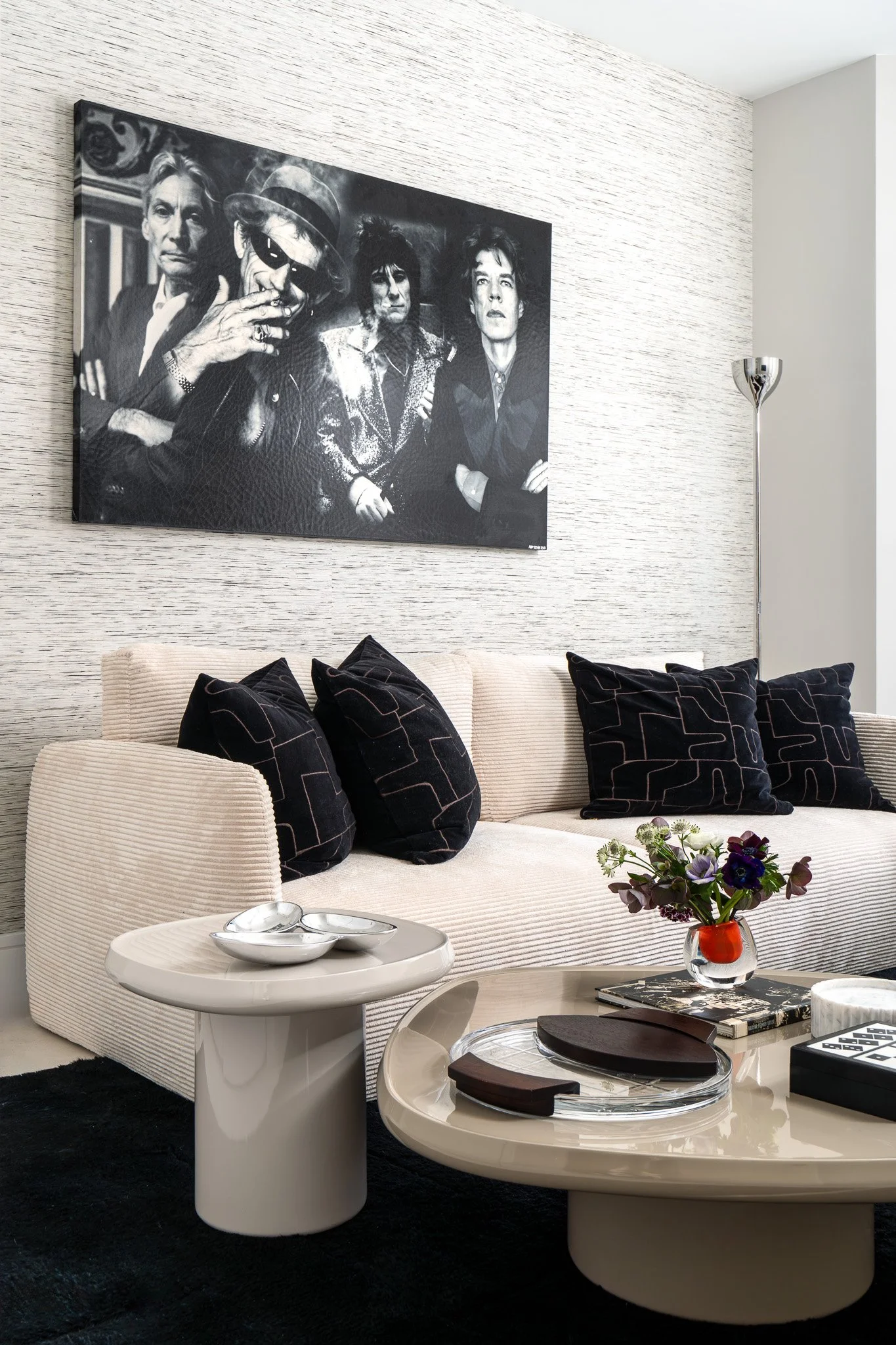 Modern living room with a beige textured sofa, black patterned cushions, a white round side table, a glossy coffee table with a tray and books, and a vase with purple and green flowers. A black and white painting hangs on the wall, and a floor lamp i