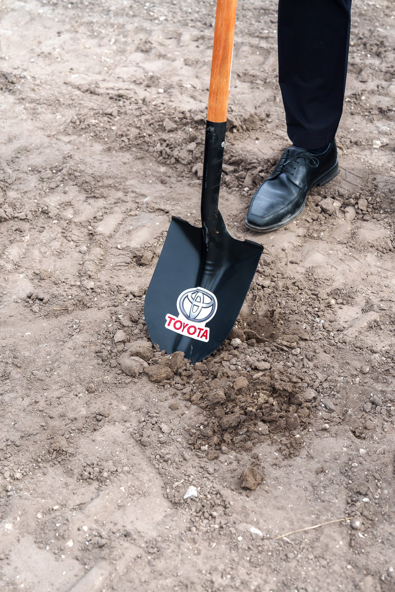 A person in black dress shoes and dark pants standing on dirt, holding a shovel with a Toyota logo on the blade.