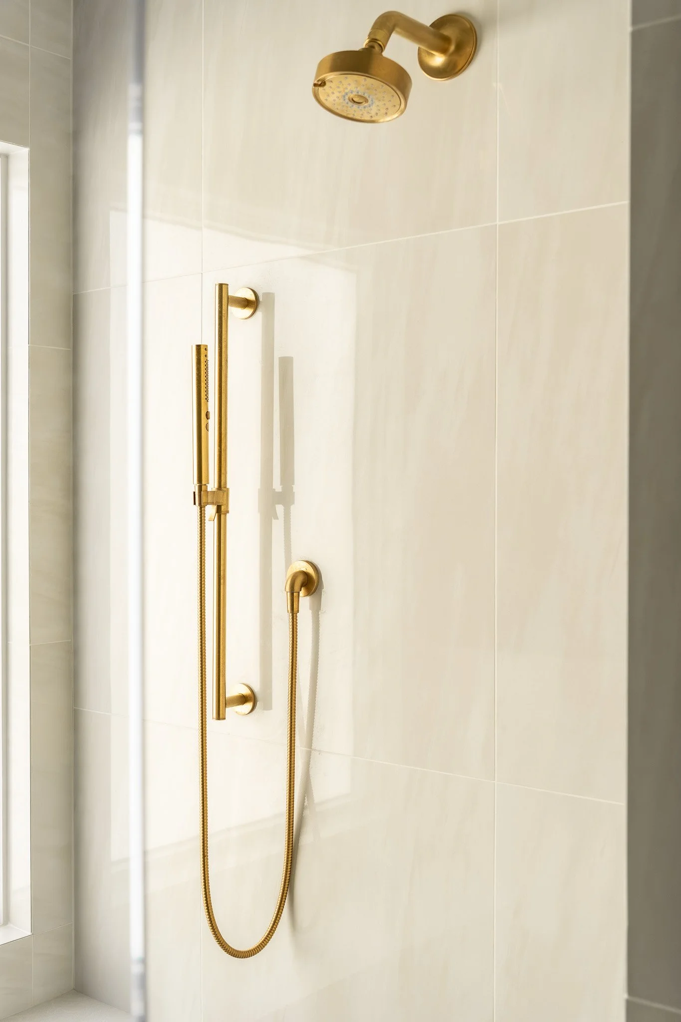 Shower with gold fixtures, including a rain showerhead, a handheld shower, and a vertical handle with a hose, installed on a light-colored tiled wall.