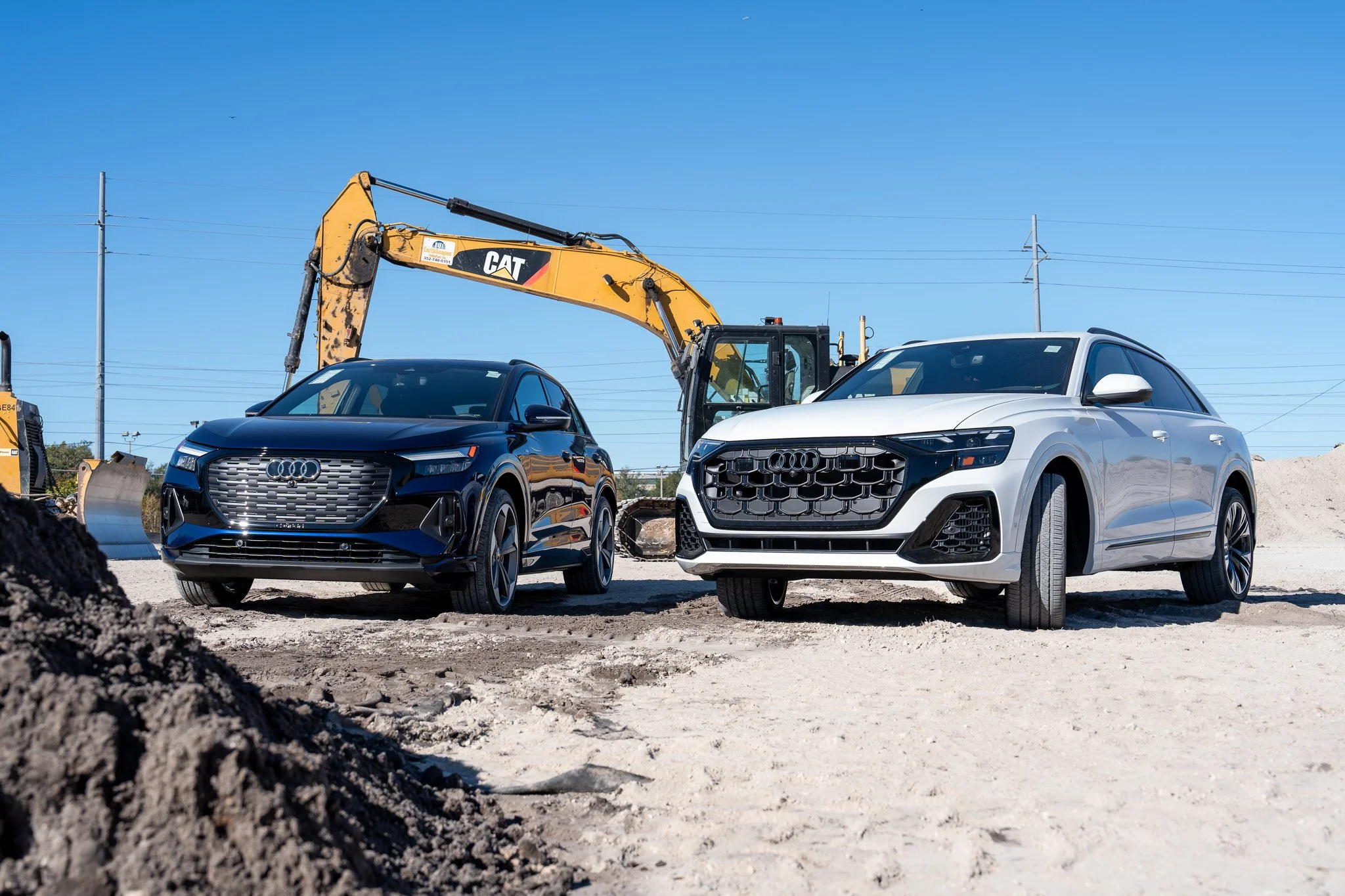 Two modern cars, one blue and one silver, parked at a construction site with a yellow excavator in the background and power lines overhead, under a clear blue sky.