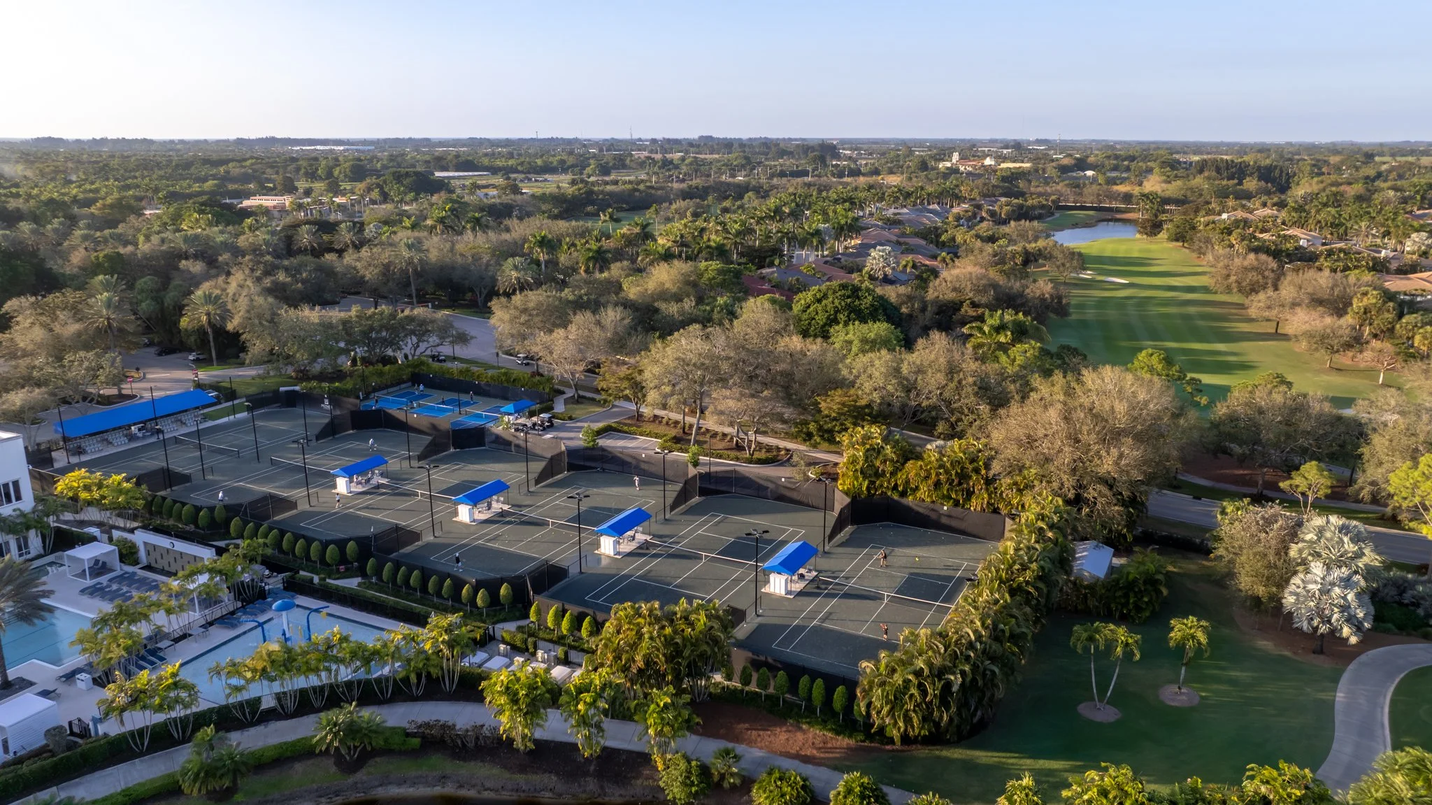 Aerial view of tennis courts, swimming pool, golf course, and surrounding trees in a lush, green landscape.