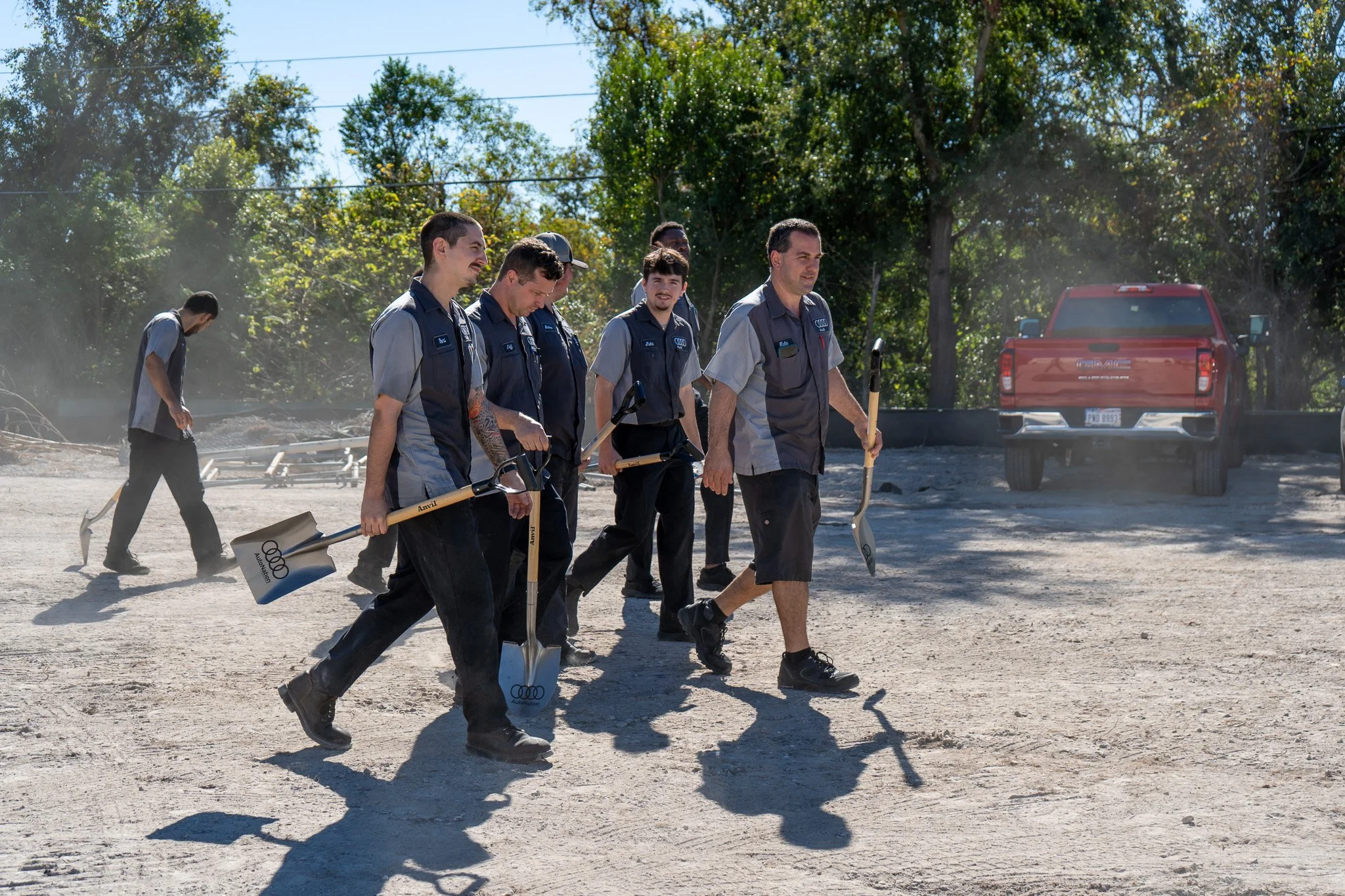 A group of seven men walking outdoors during the daytime. They are dressed in work uniforms and carrying shovels, with a red truck in the background. The scene appears to be a worksite or community project.
