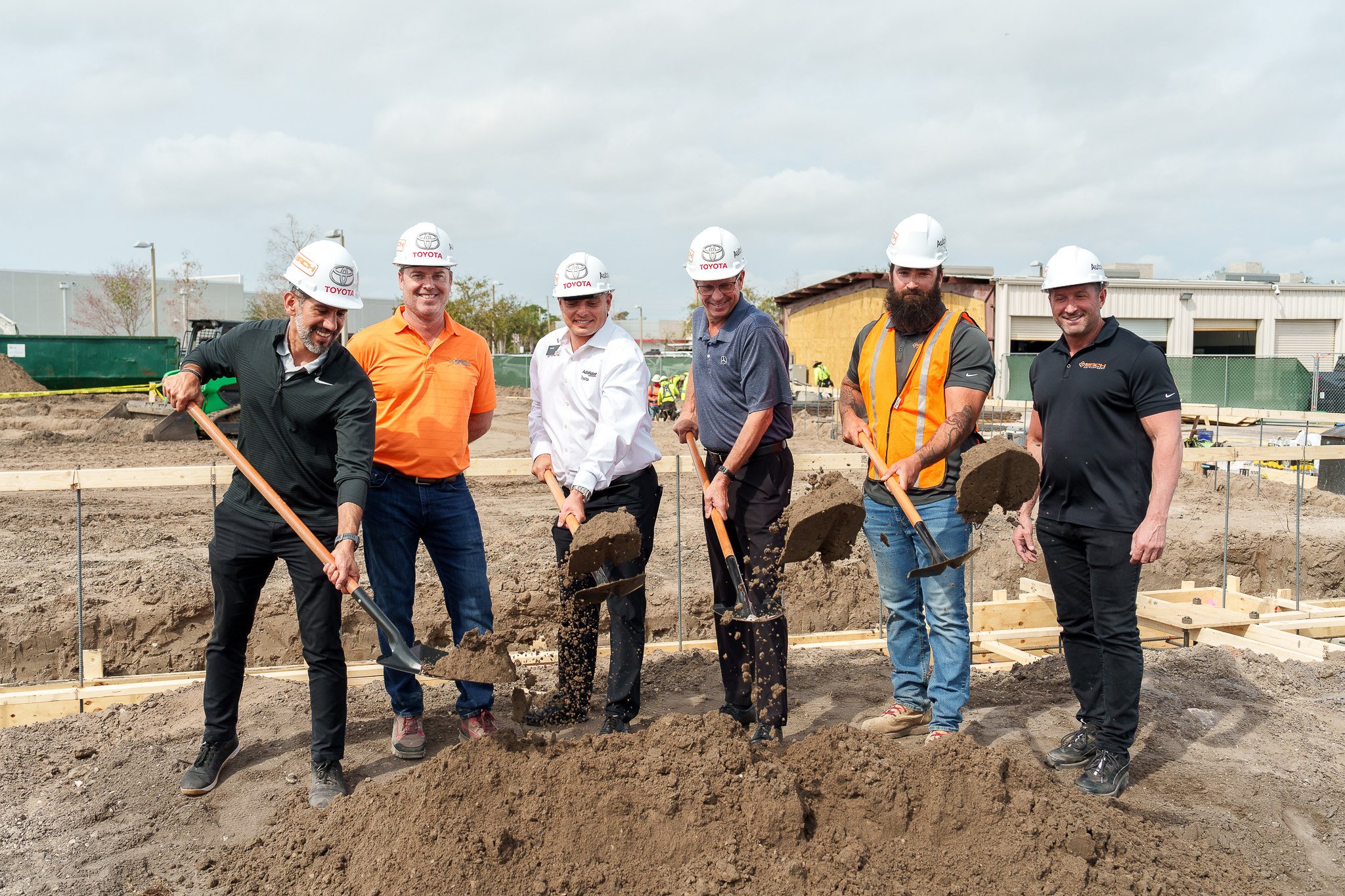 Five men in construction helmets and work attire are shoveling dirt at a construction site, with other workers and buildings in the background.