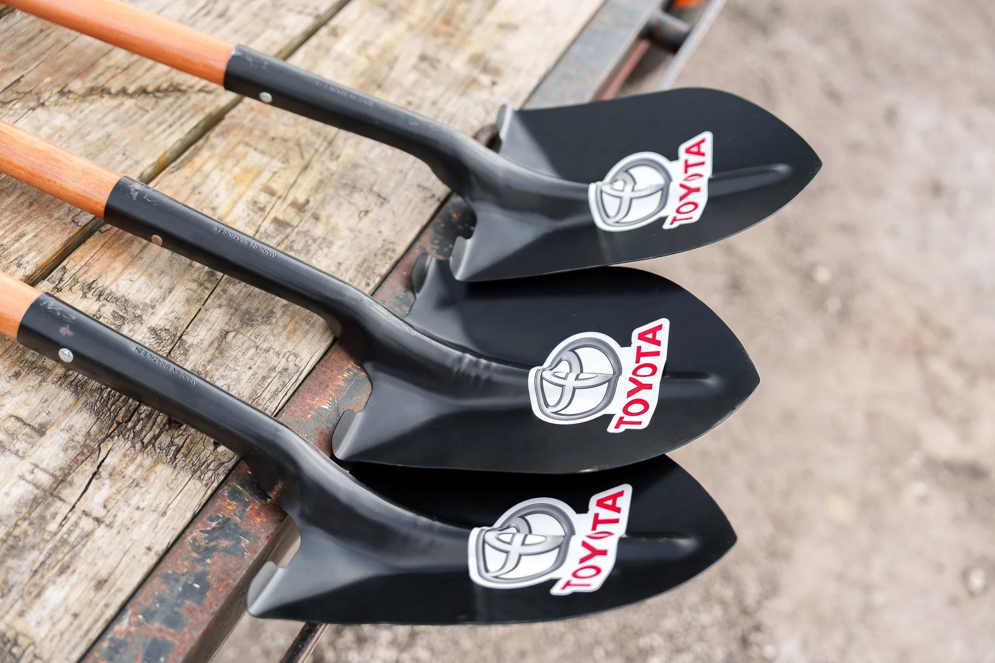 A row of three small shovels with black blades and wooden handles, neatly placed on a weathered wooden surface outdoors, each displaying a Toyota logo sticker.