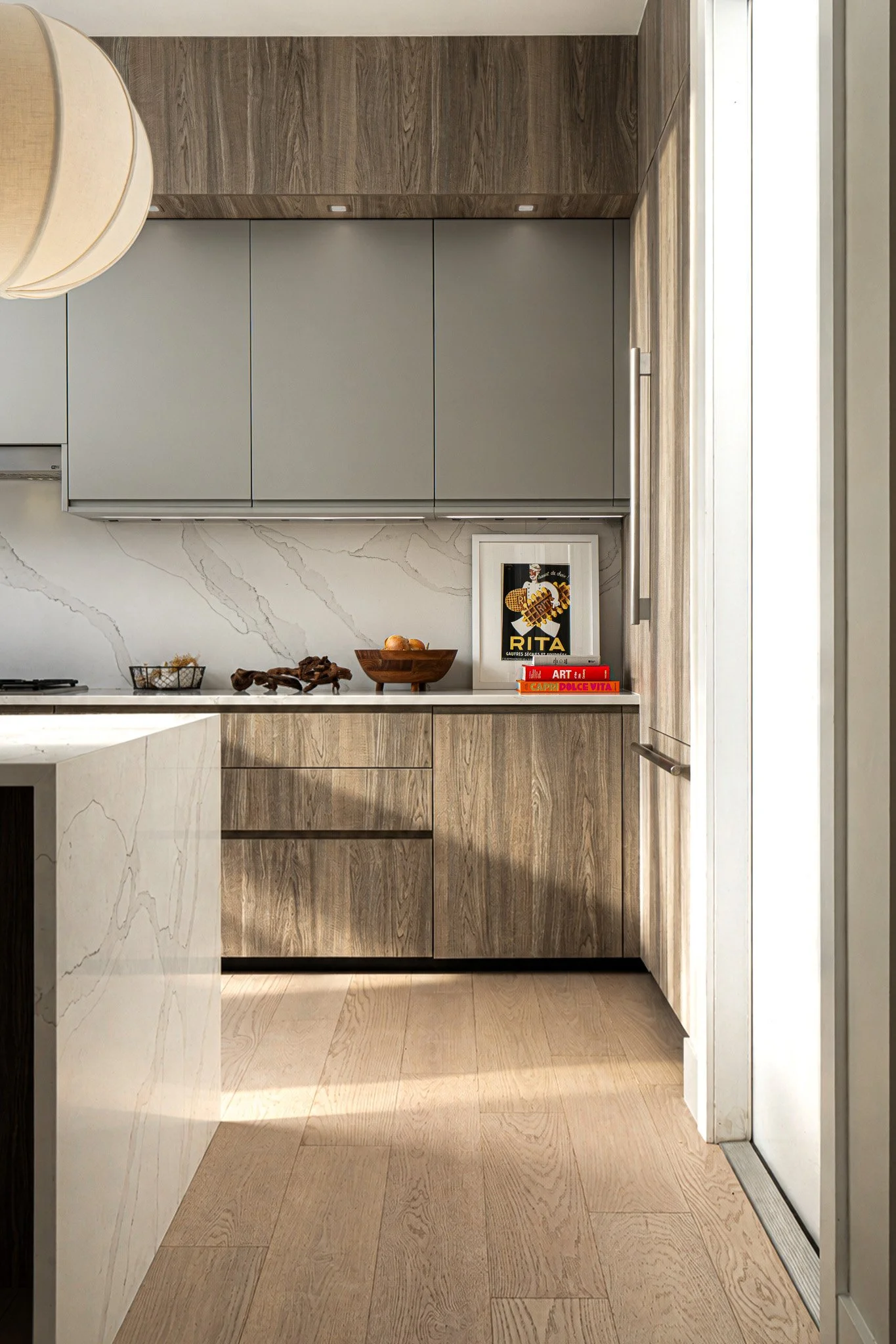 Modern kitchen with light wood and gray cabinetry, marble backsplash, and decorative items including books, framed art, and bowls on the countertop.