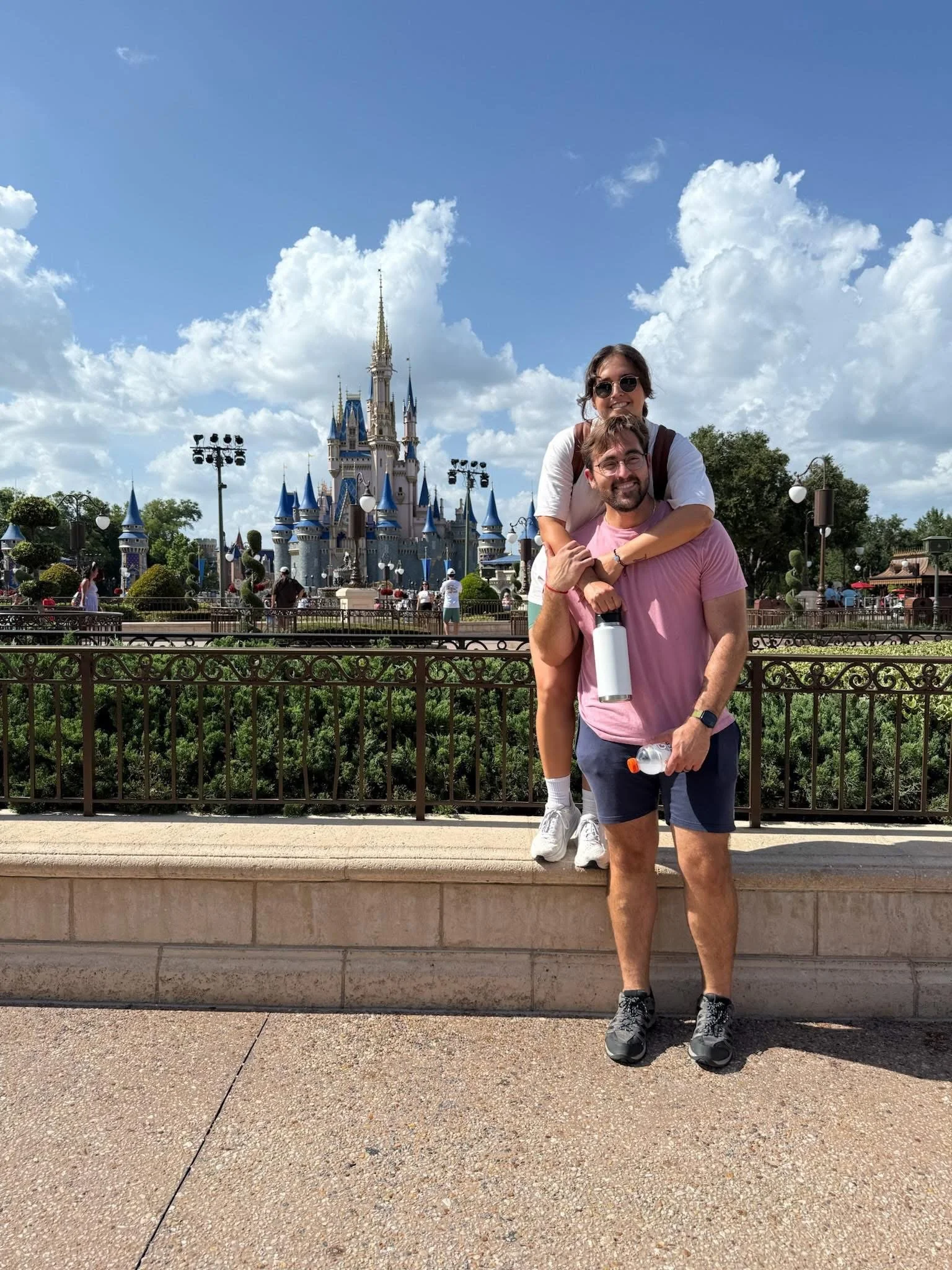 A couple posing in front of Cinderella's Castle at Disney World theme park, with the woman sitting on the man's shoulders. The sky is blue with some clouds, and there are people and greenery in the background.