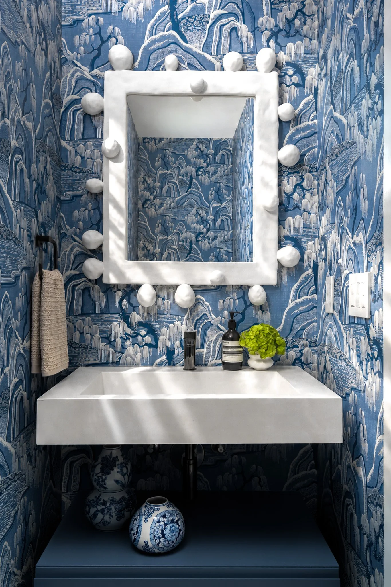 A bathroom with blue and white patterned wallpaper and ceramics, including a white floating sink, a mirror surrounded by white spherical lights, a hand towel, and decorative vases.