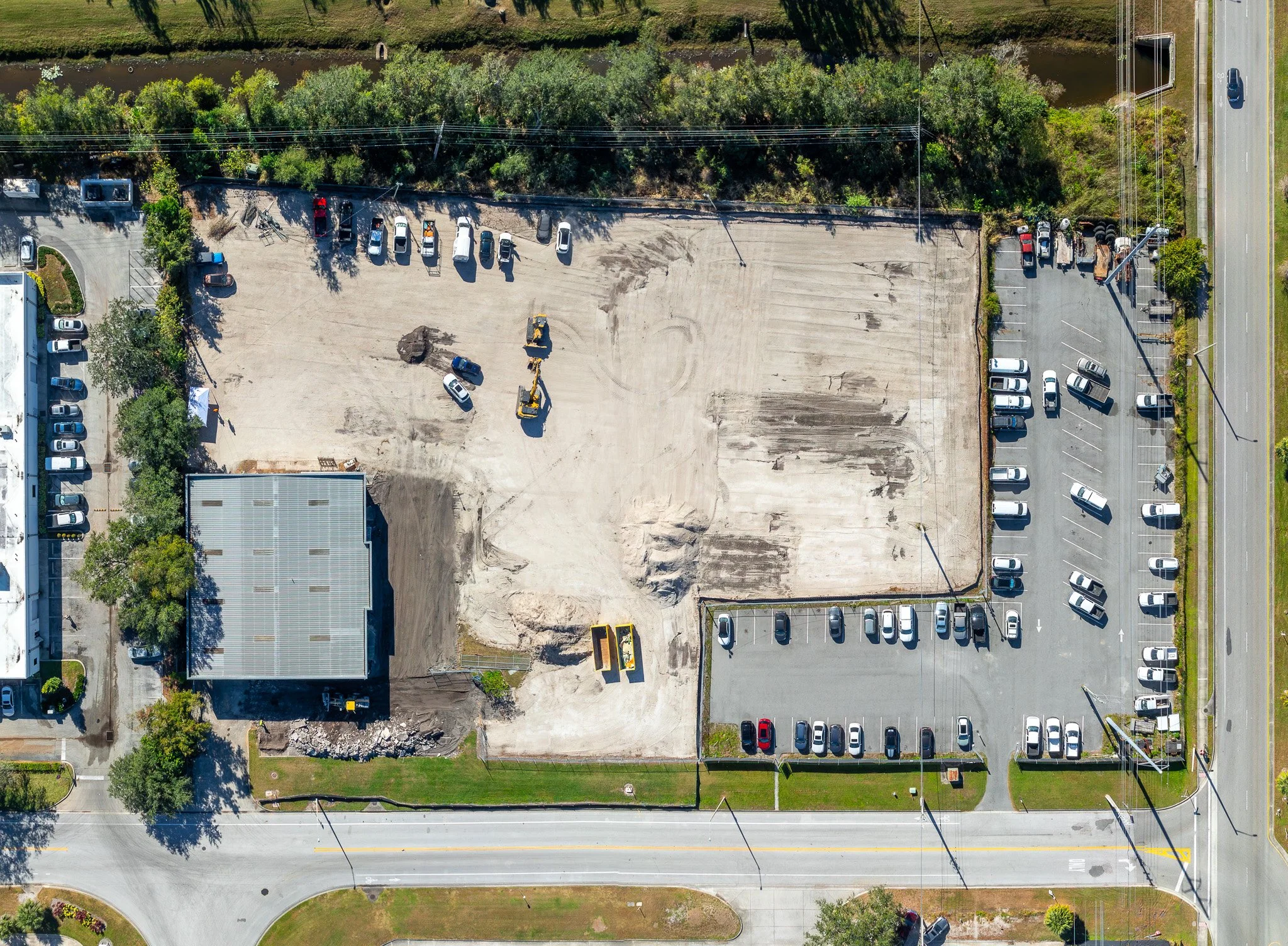 An aerial view of a construction site with some parked cars, construction equipment, and surrounding trees and roads.