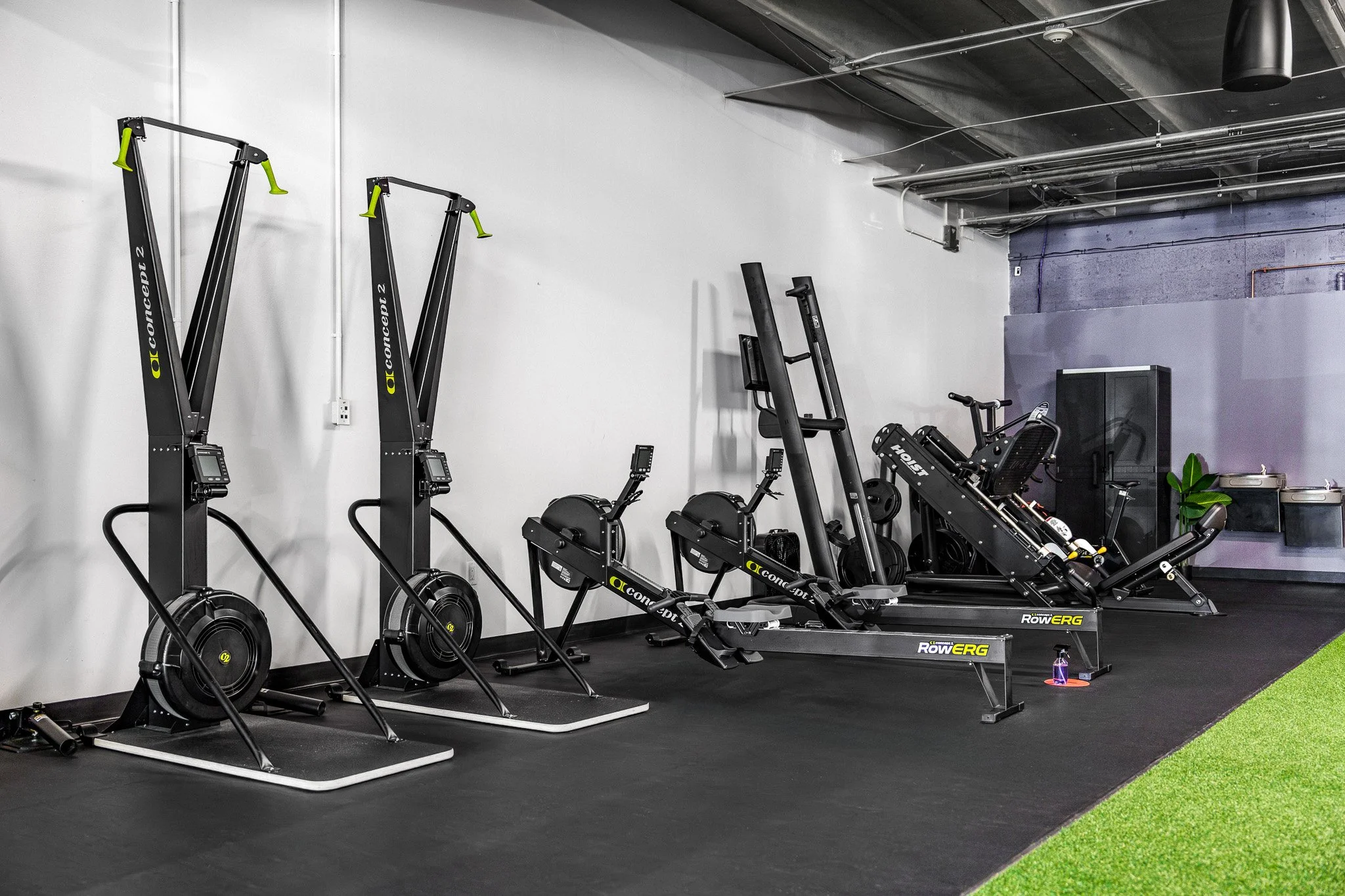 A gym with multiple rowing machines and exercise equipment arranged in a row, with a black rubber floor and a white wall behind.
