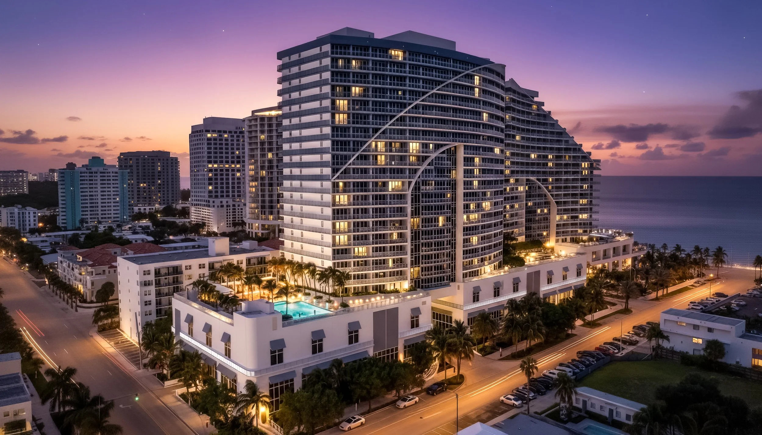 High-rise residential building near the ocean at dusk, with lights on in the apartments and a pool area with palm trees on the ground level.