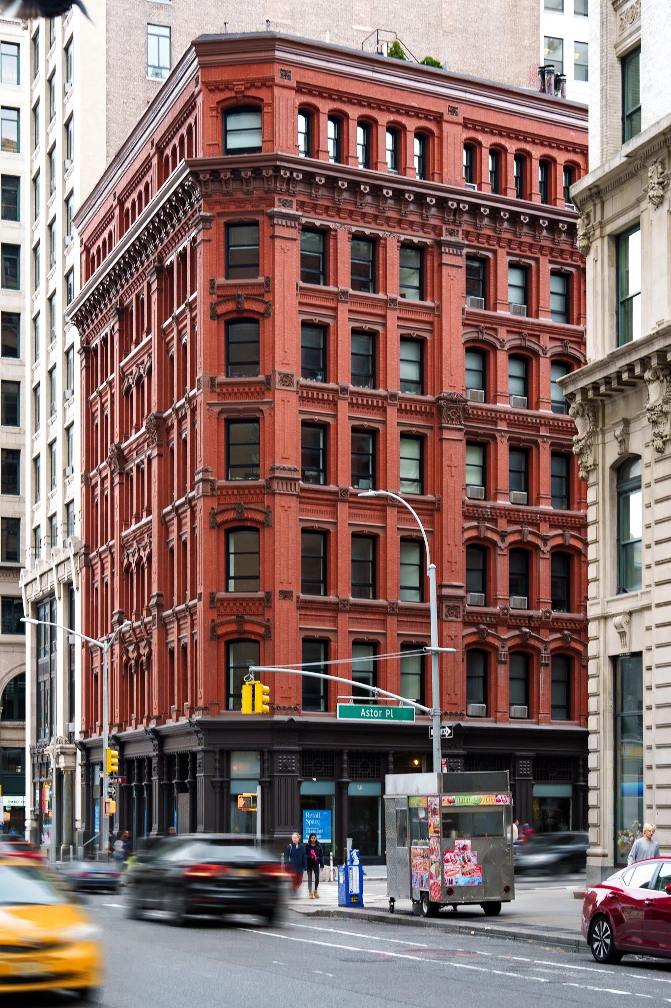 Urban city street with a red brick building at the corner, busy traffic, pedestrian crossing, street sign reading 'Astor Pl', and a food cart on the sidewalk.