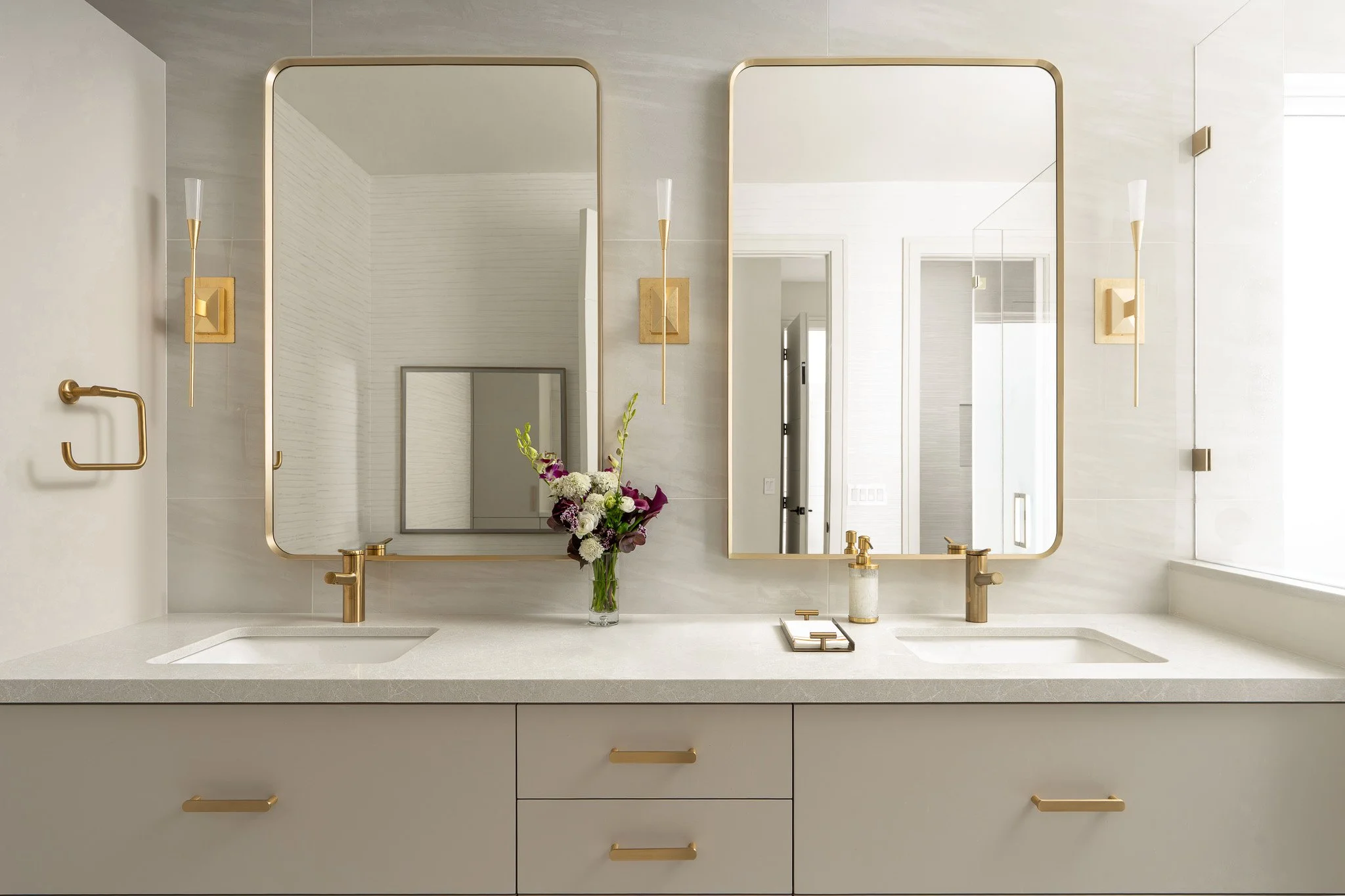 Modern bathroom vanity with two large mirrors, gold fixtures, and a white countertop with a vase of flowers.