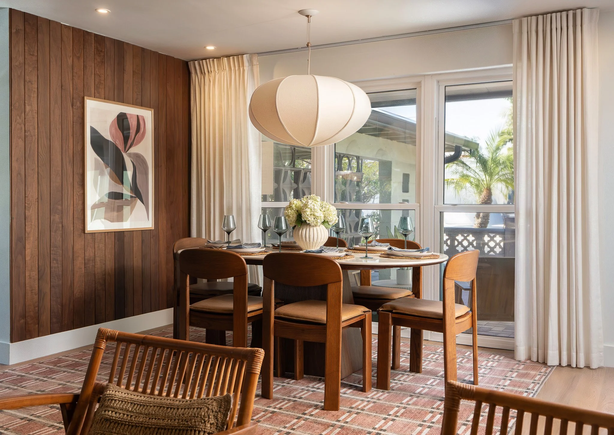 Dining room with wooden wall paneling, round dining table with six wooden chairs, white curtain, large window showing a palm tree outside, and a modern white pendant light hanging above the table.