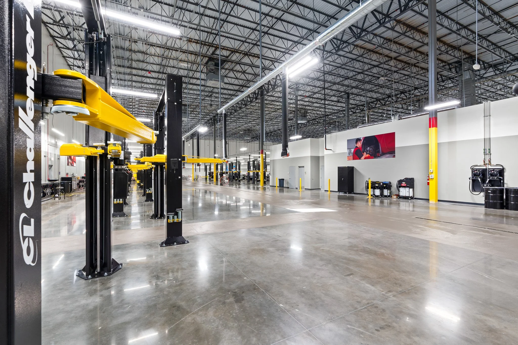 Empty auto repair workshop with yellow vehicle lifts, a large digital screen showing a mechanic working on a car, and polished concrete floors.
