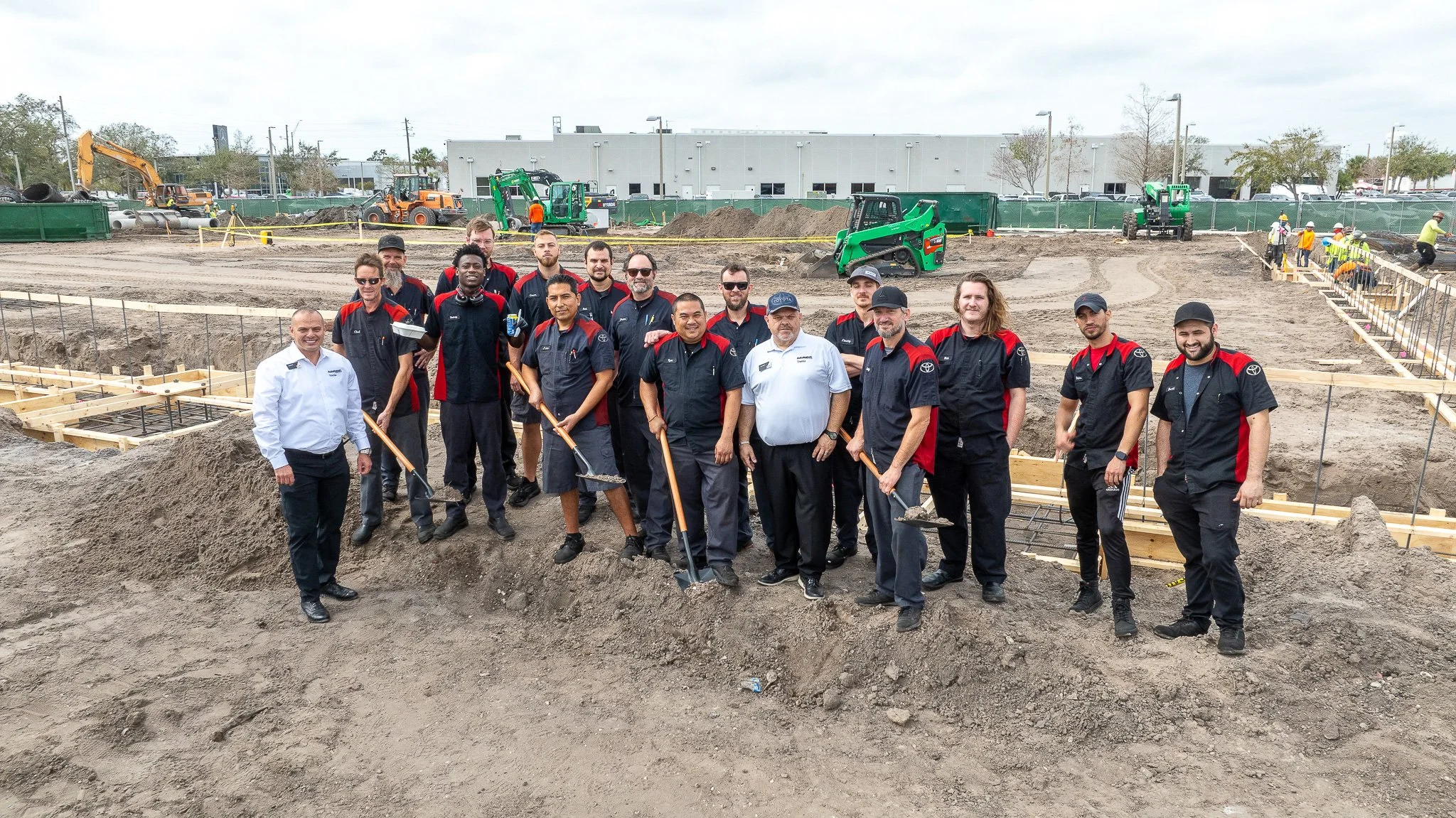 Group of construction workers and employees posing for a photo at a construction site with machinery and dug-out trenches in the background.