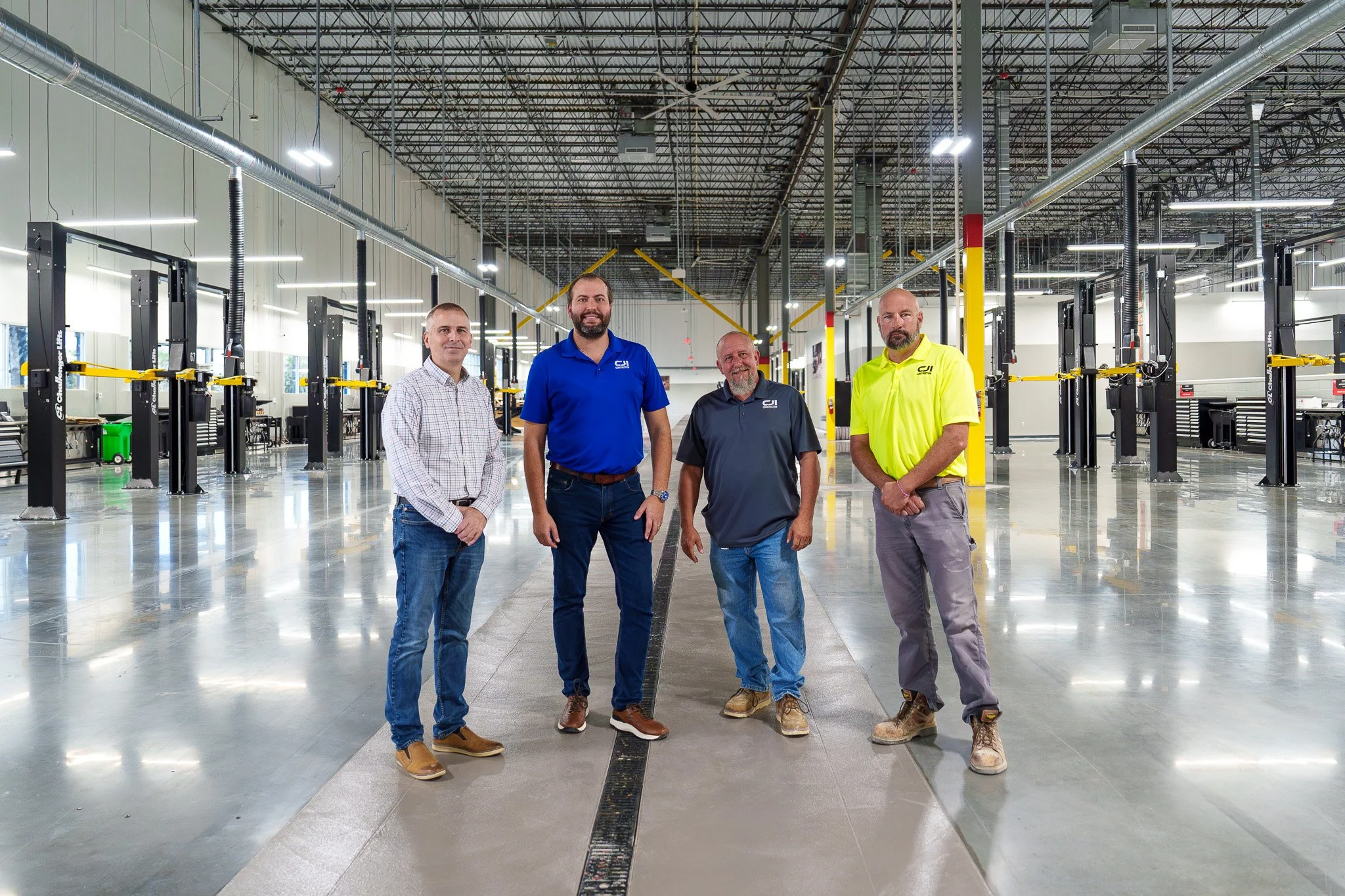 Four men standing in a large, industrial-style gym or warehouse with high ceilings, bright lighting, and fitness equipment in the background.