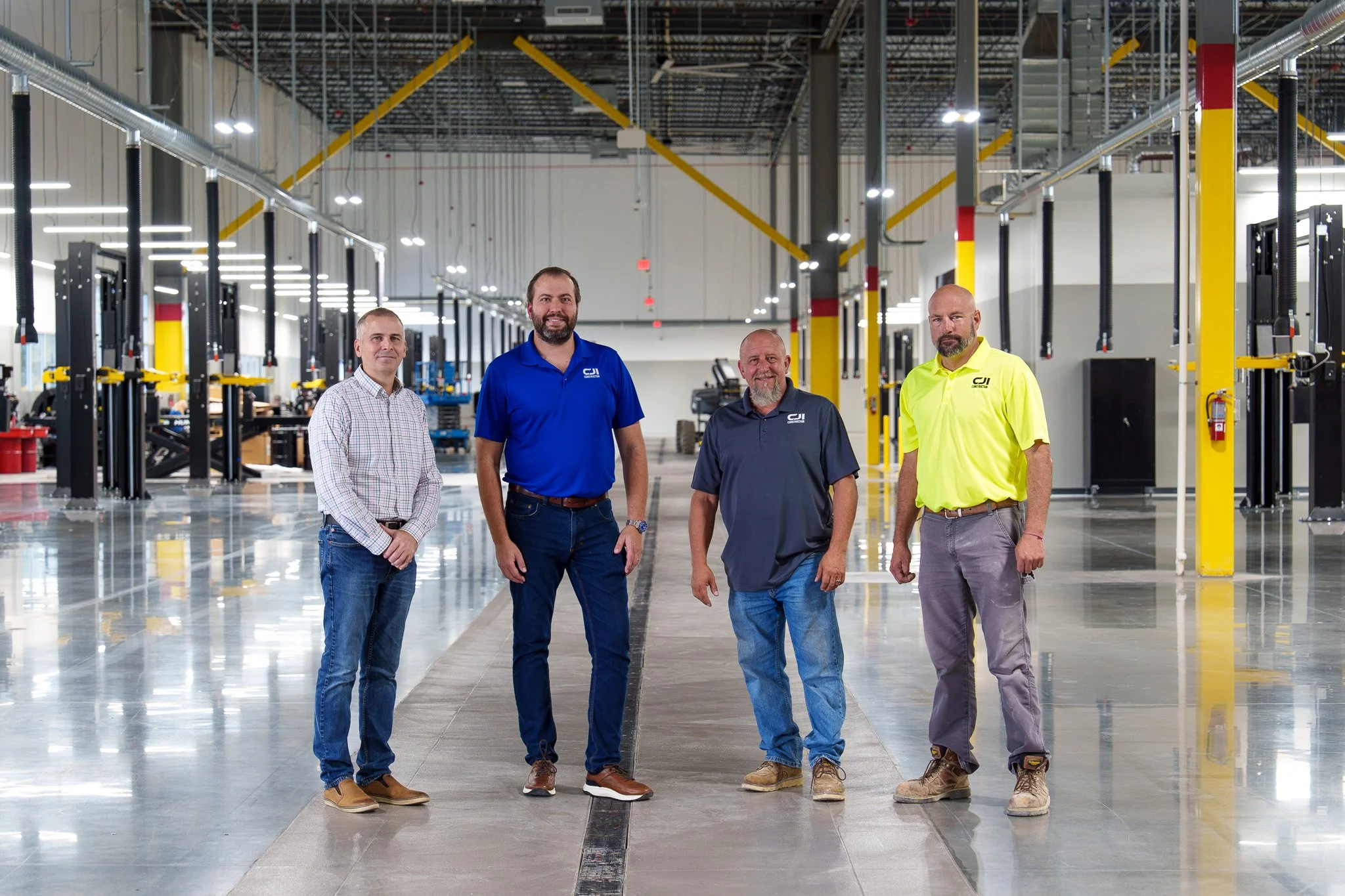 Four men standing in a large, industrial workshop or factory, with manufacturing equipment and bright lighting overhead.