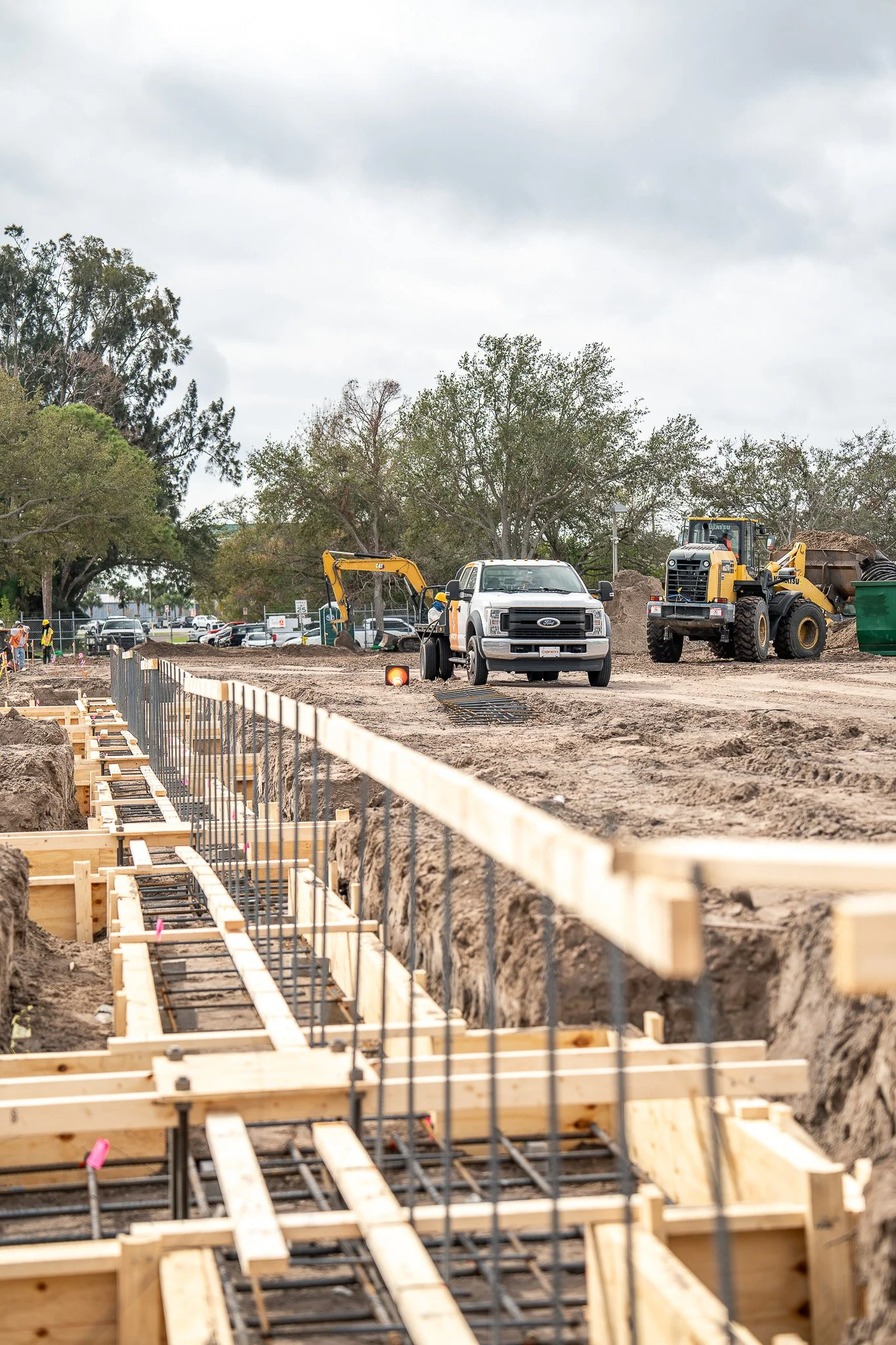 Construction site with wooden and metal framework for a building foundation, heavy machinery, and workers in safety gear under an overcast sky.