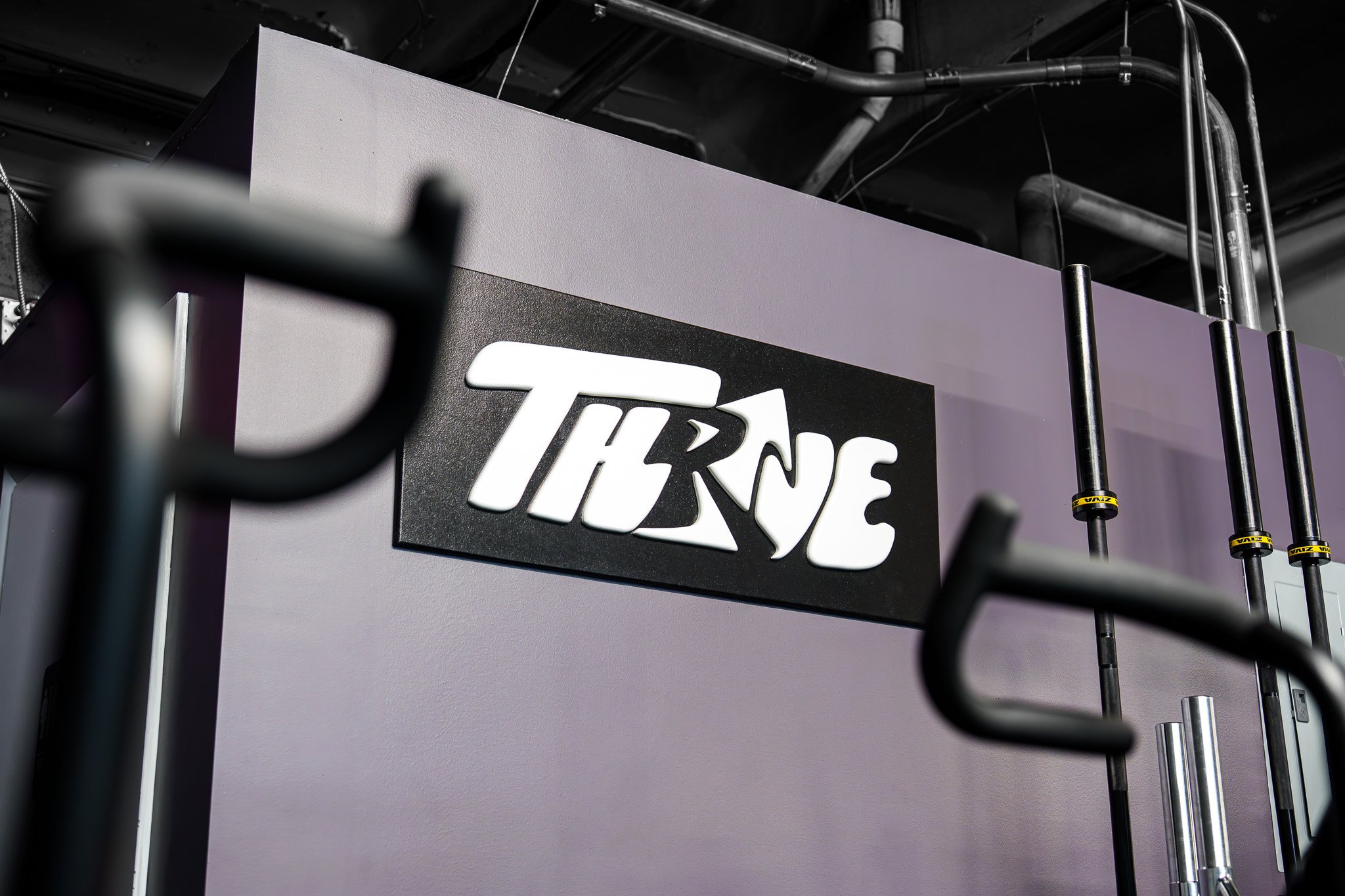 Close-up of a sign with the partial logo and text 'Thrive' on a gray wall, with industrial pipes and yellow and black Valpar fittings in the background.