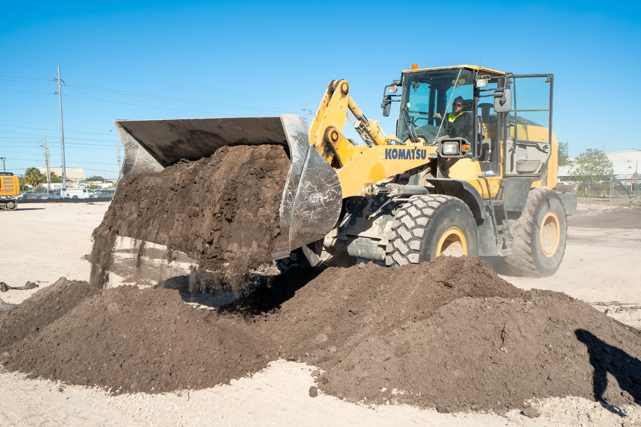 A yellow Komatsu front loader moving dirt at a construction site, with power lines and buildings in the background under a clear blue sky.