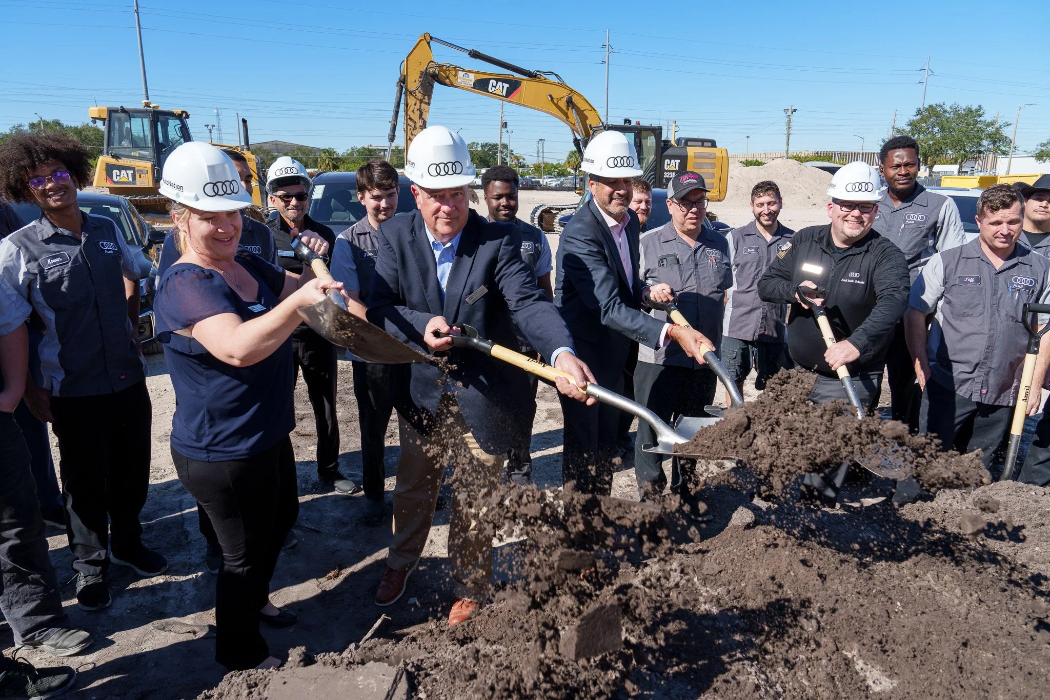 Group of people, including some wearing construction helmets, gathered at a construction site while shoveling dirt, with large construction machinery and parked cars in the background.