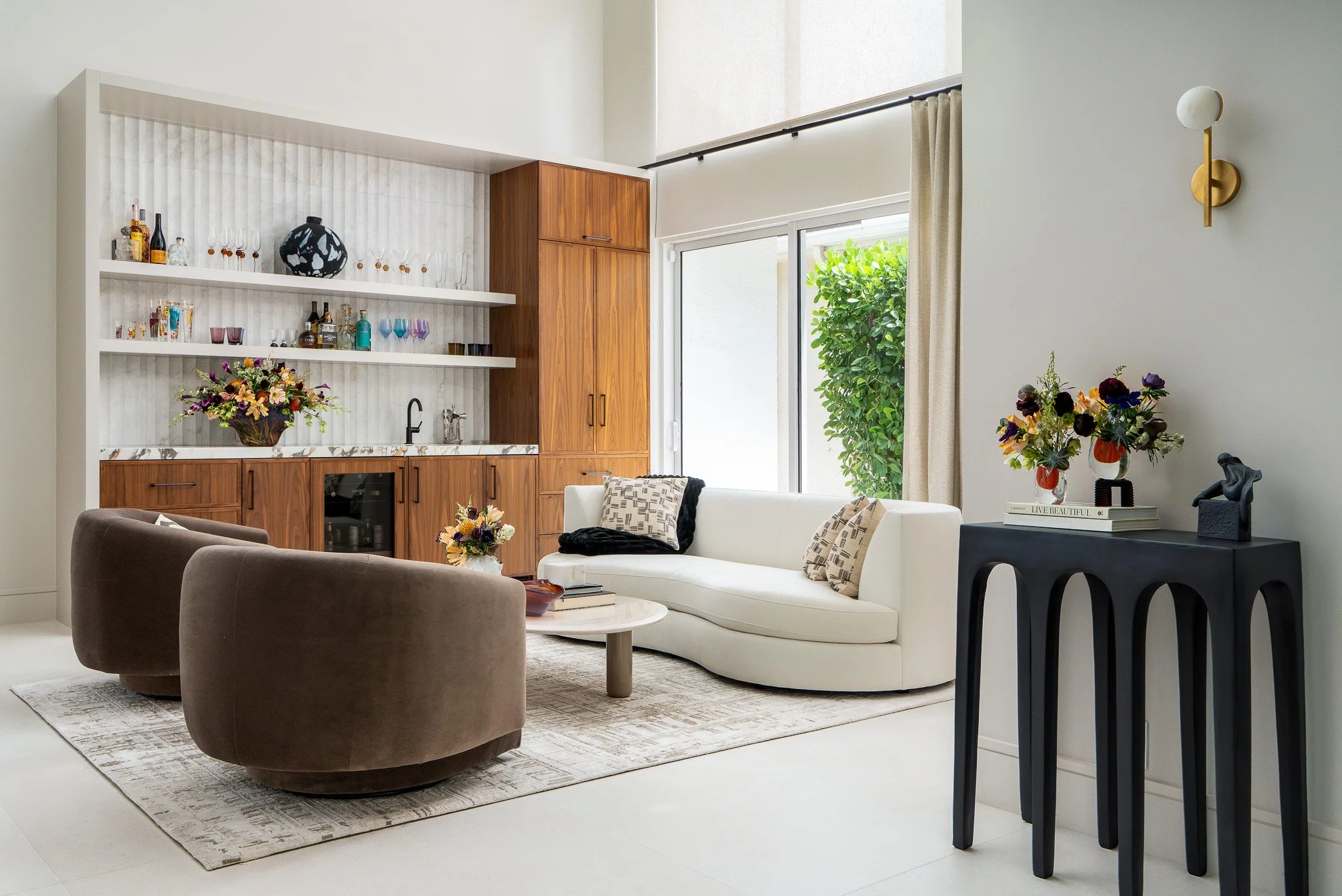 A modern living room with a white curved sofa, a round marble-top coffee table, and a brown armchair. Decor includes two vases with flowers, stylish books, and a black sculpture. The wall has a built-in wooden cabinet with open shelves displaying gla