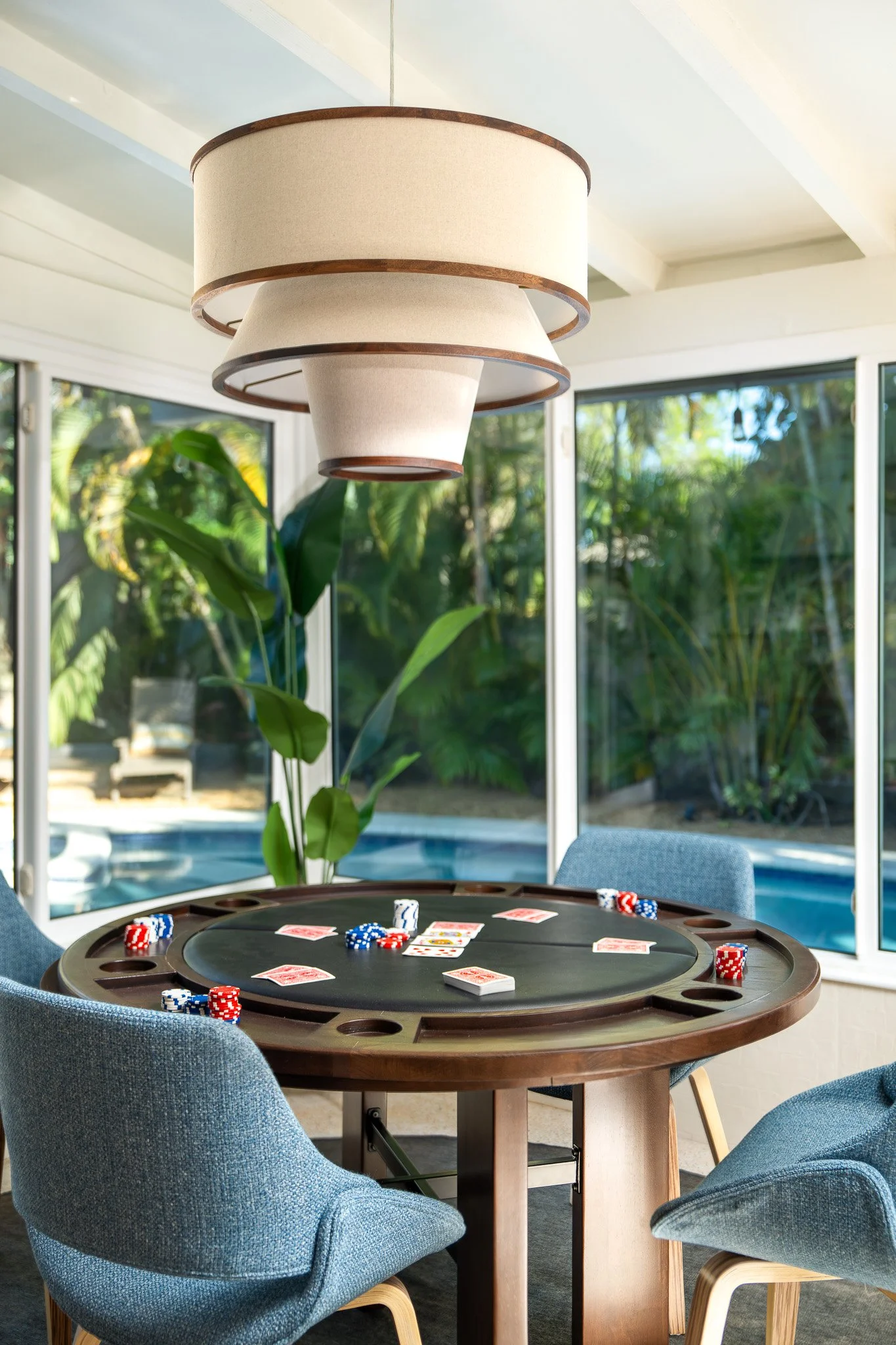 A round poker table with chips, cards, and player chairs inside a bright room with large windows showing green outdoor foliage.
