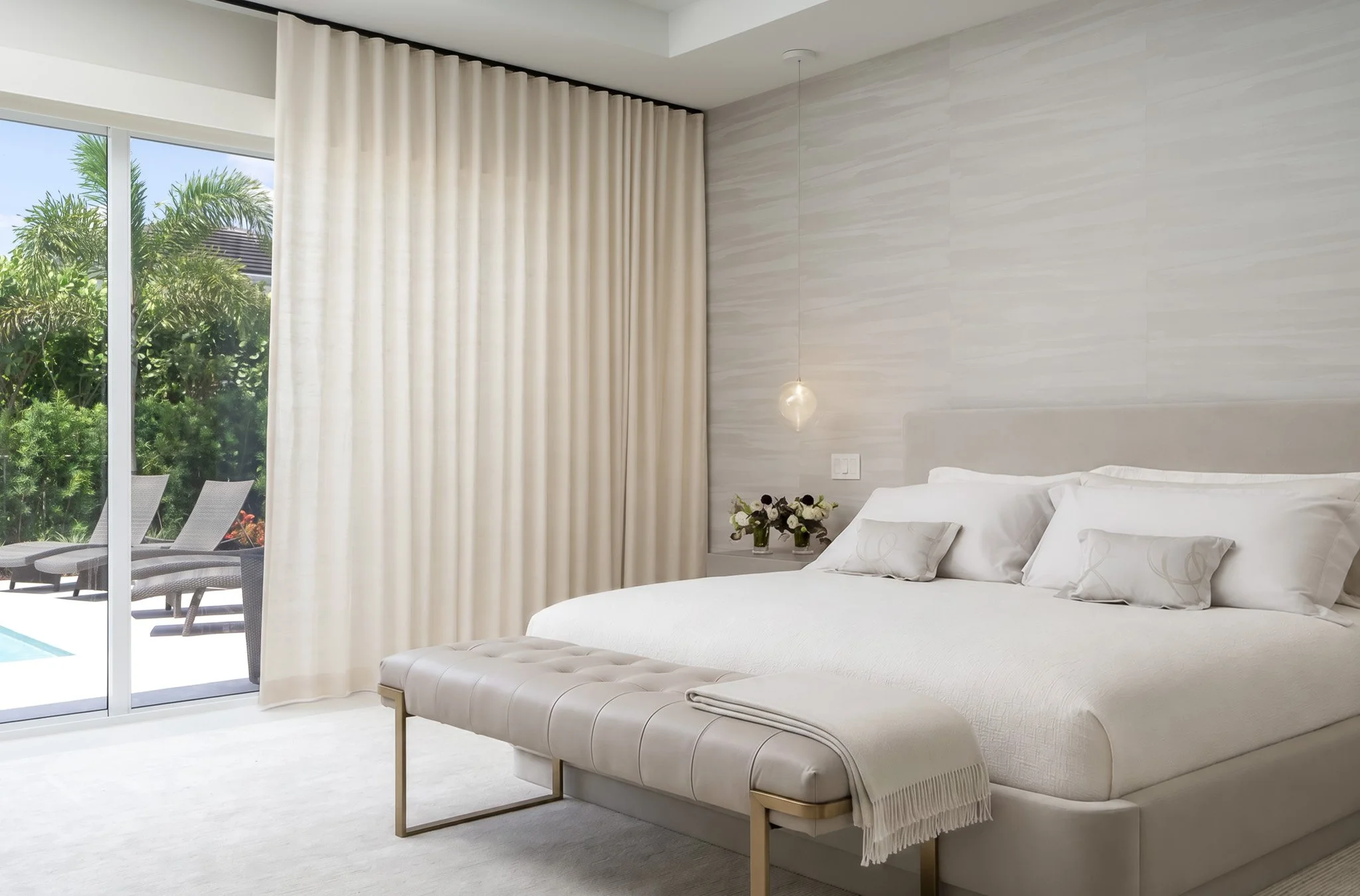 A bedroom with a large bed with white bedding and pillows, a small bedside table with a flower arrangement, a pendant light, and large sliding glass doors leading to an outdoor patio with chairs and trees.