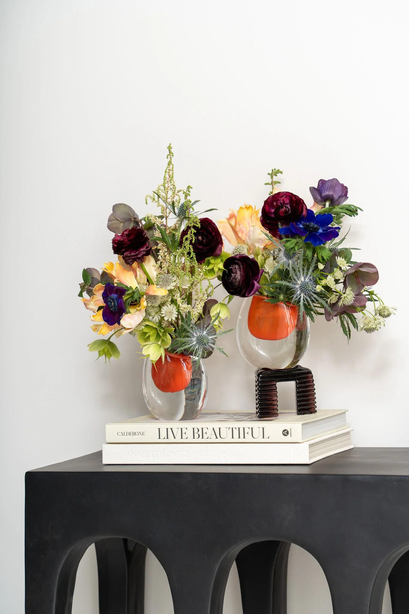 Two vases with colorful flowers on top of a white book titled 'LIVE BEAUTIFUL' placed on a black surface, with a black decorative object behind the vases. The background is plain white.