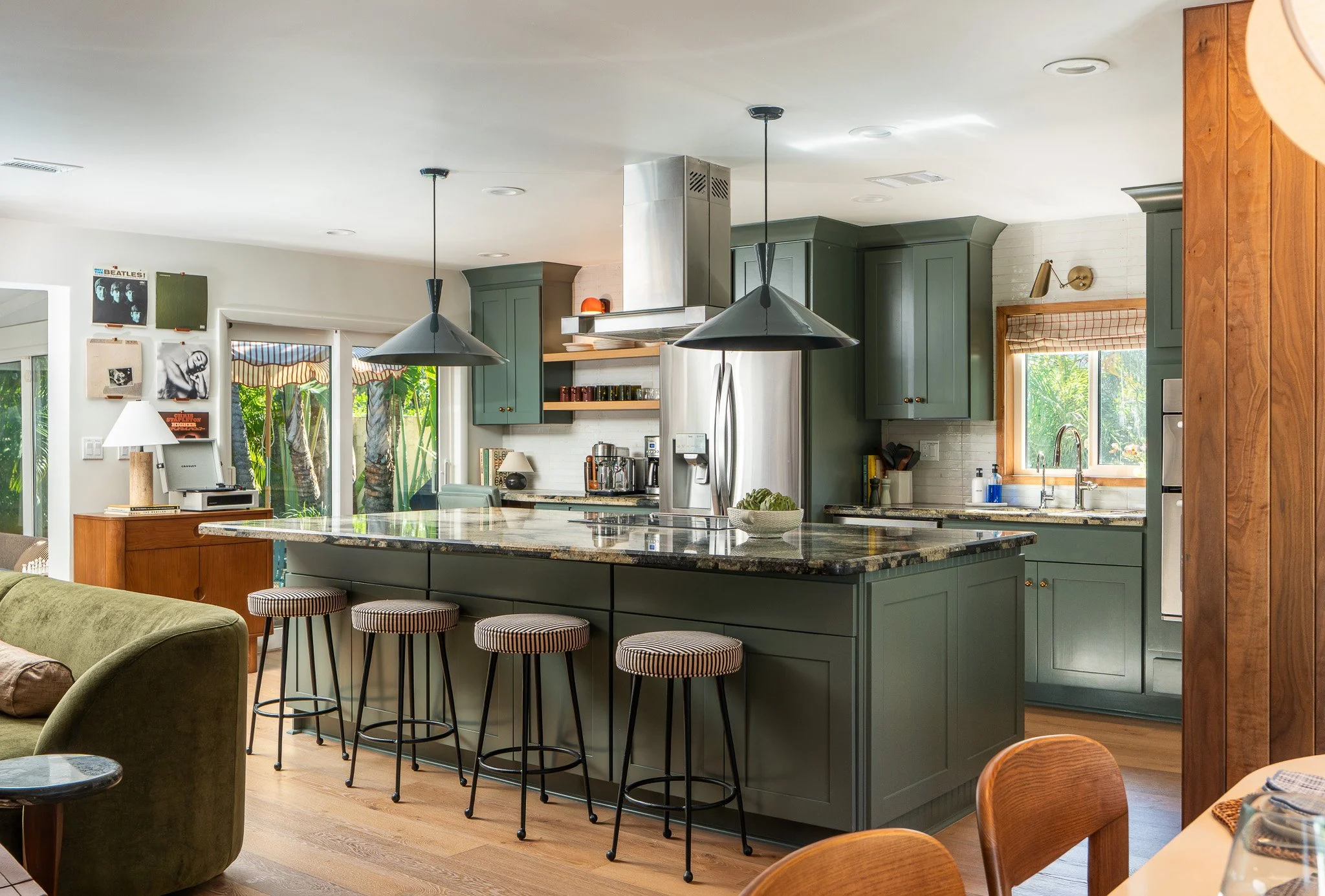 A kitchen with green cabinetry, a large granite island with bar stools, stainless steel refrigerator, double sink, pendant lights, and windows with green foliage outside.