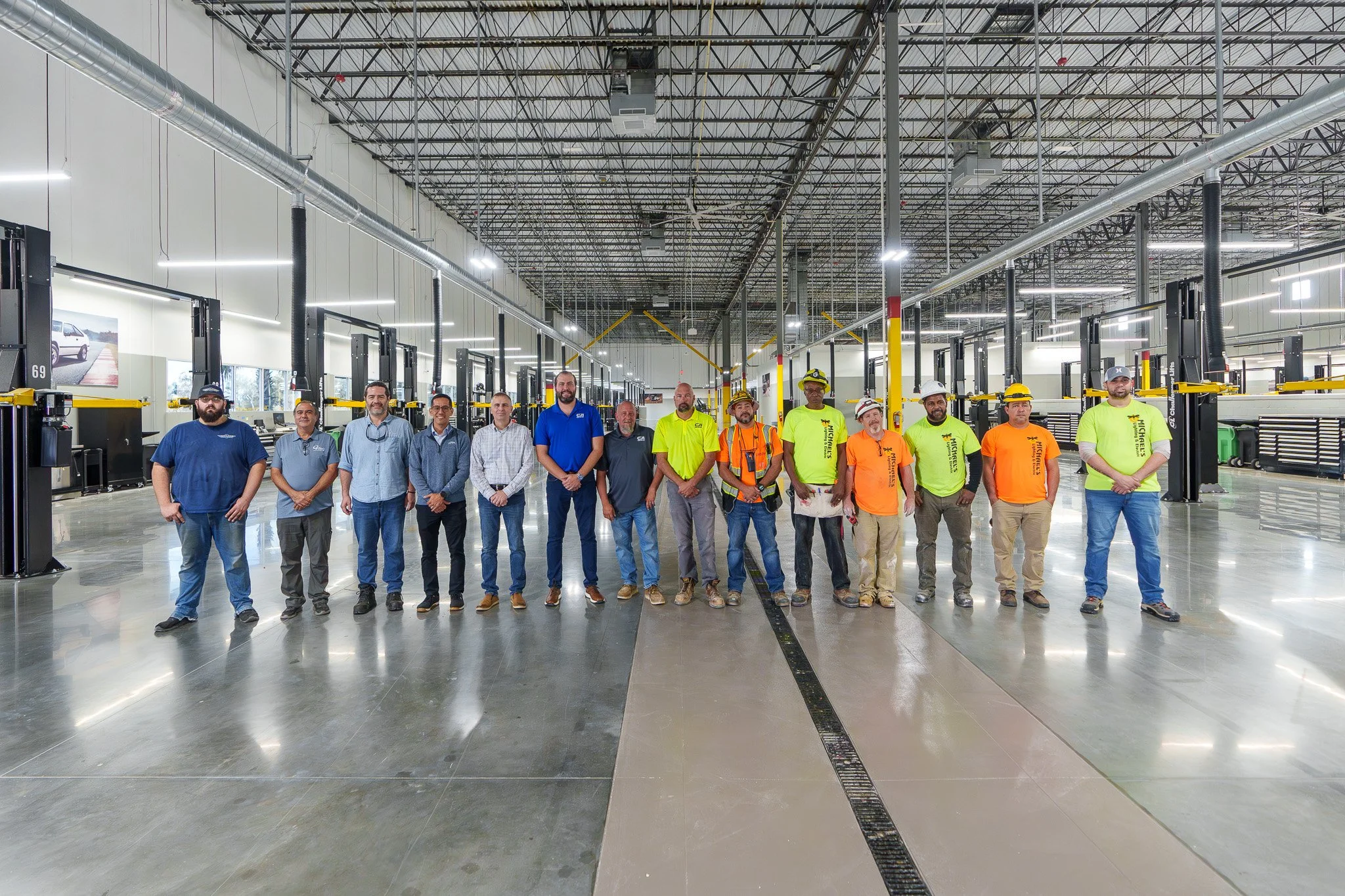 Group of people standing inside a large industrial or warehouse facility with high ceilings, exposed ductwork, and rows of equipment or workstations.
