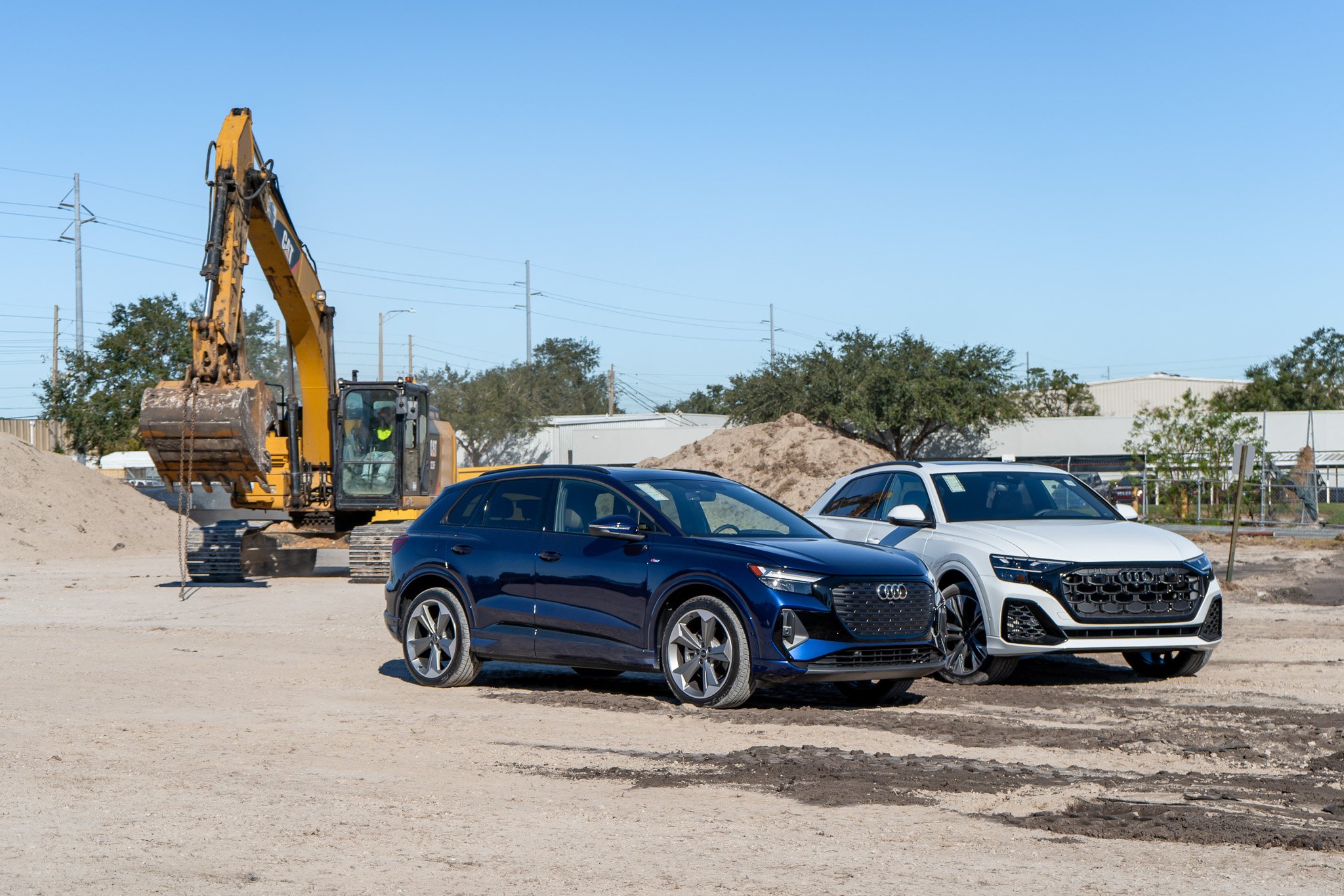 Construction site with a yellow excavator and two parked cars, one black and one white, under a clear blue sky.