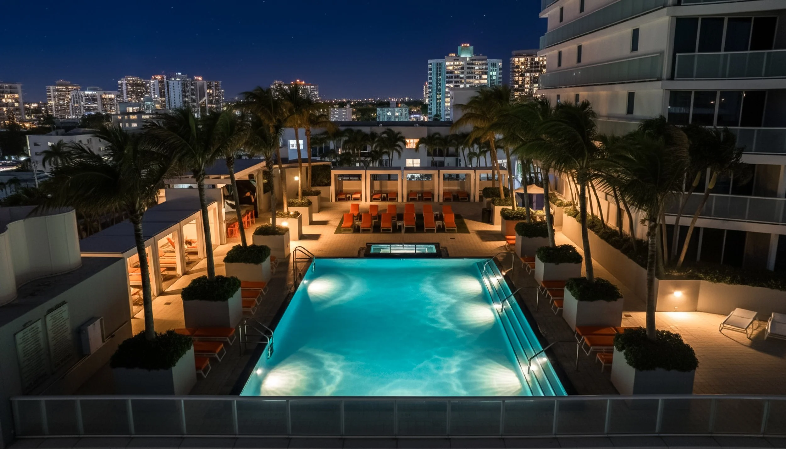 Nighttime view of a rooftop pool area with lounge chairs, palm trees, and city skyline in the background.
