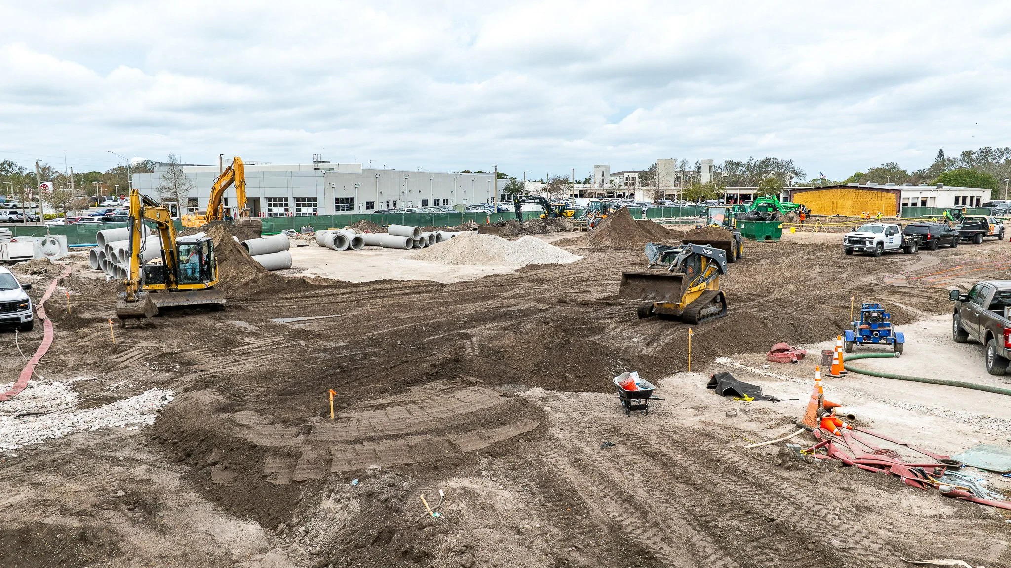 Construction site with excavators, dirt piles, concrete pipes, and vehicles under cloudy sky.