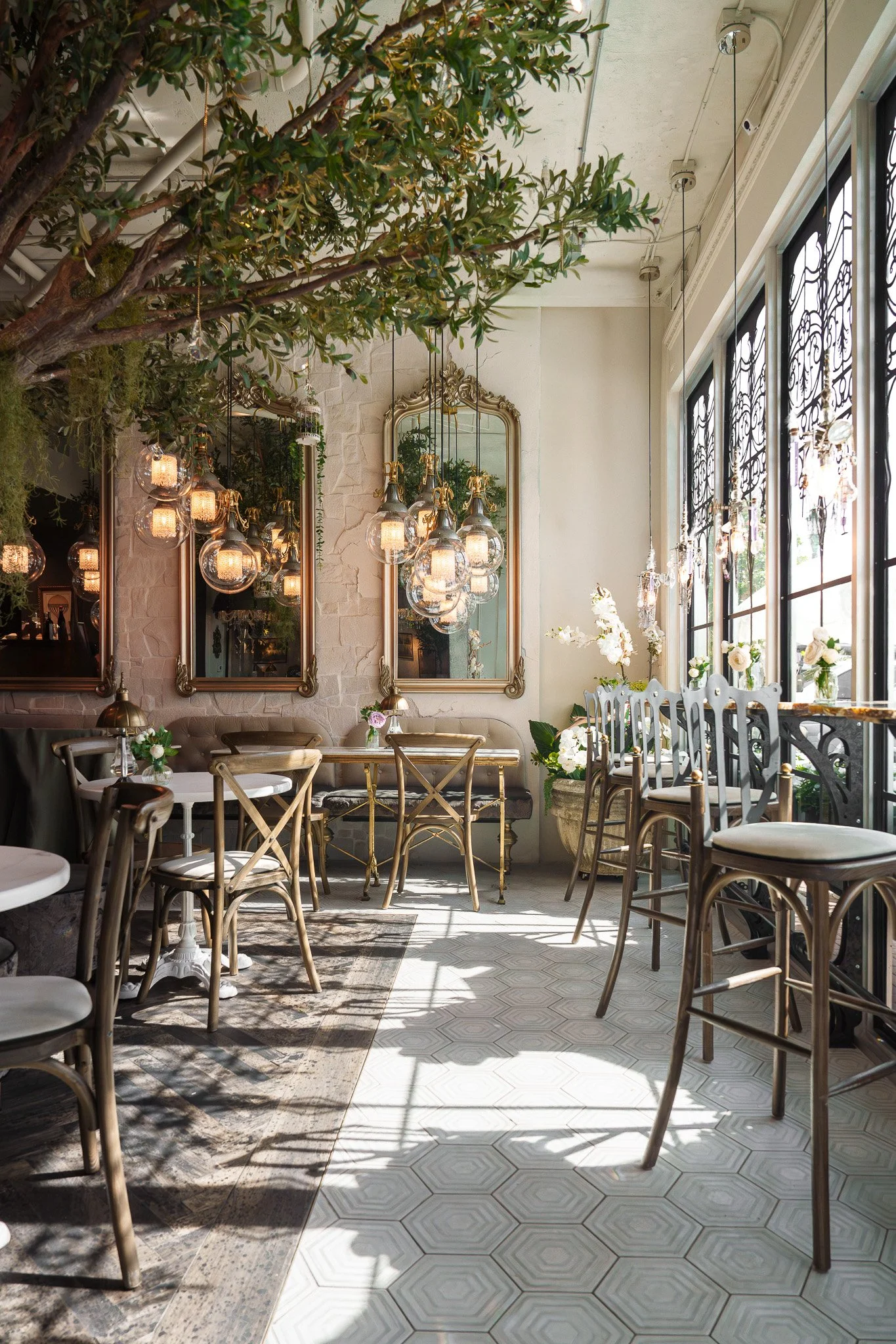 Interior of a restaurant or café with large windows, hanging light fixtures, a large green tree, and decorated tables with flowers. Mirrors hang on a white brick wall.