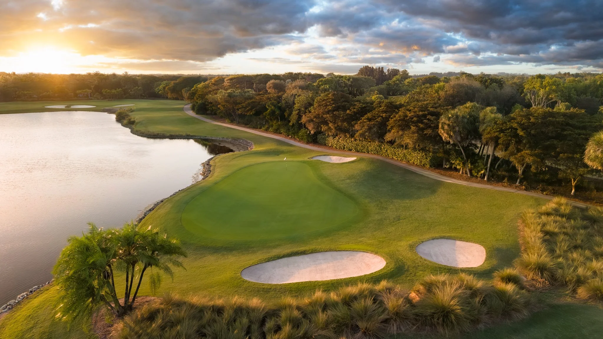 A golf course with a green, sand bunkers, and a water hazard, with trees in the background and a cloudy sky, illuminated by golden sunlight.