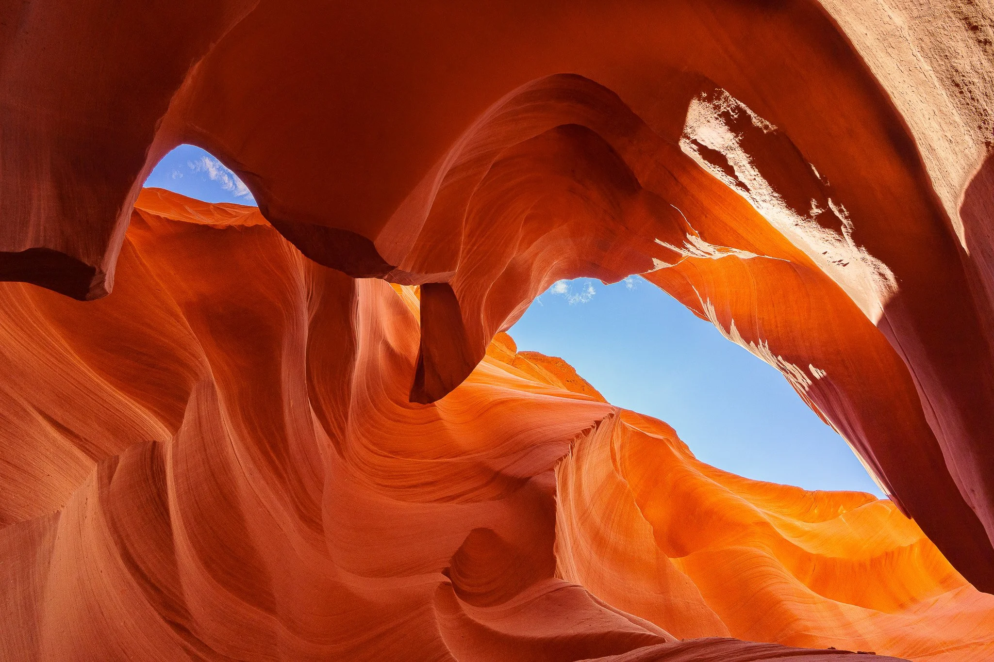 View of a narrow slot canyon with smooth, curved sandstone walls in shades of orange and red, with a small opening showing a blue sky and white clouds above.