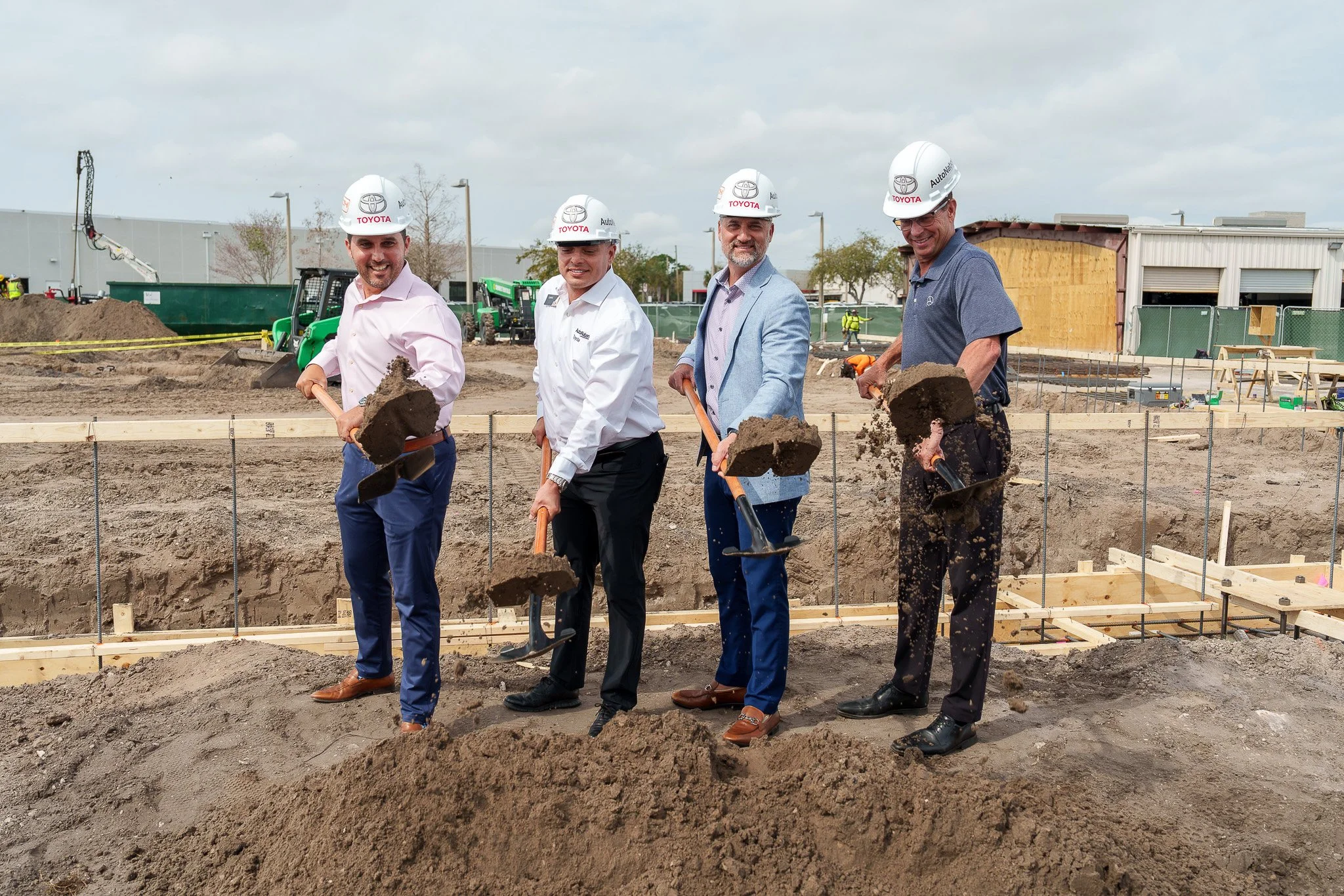 Four men in dress shirts and suit jackets wearing safety helmets with Toyota logos, shoveling dirt on a construction site, with machinery and partially built structures in the background.