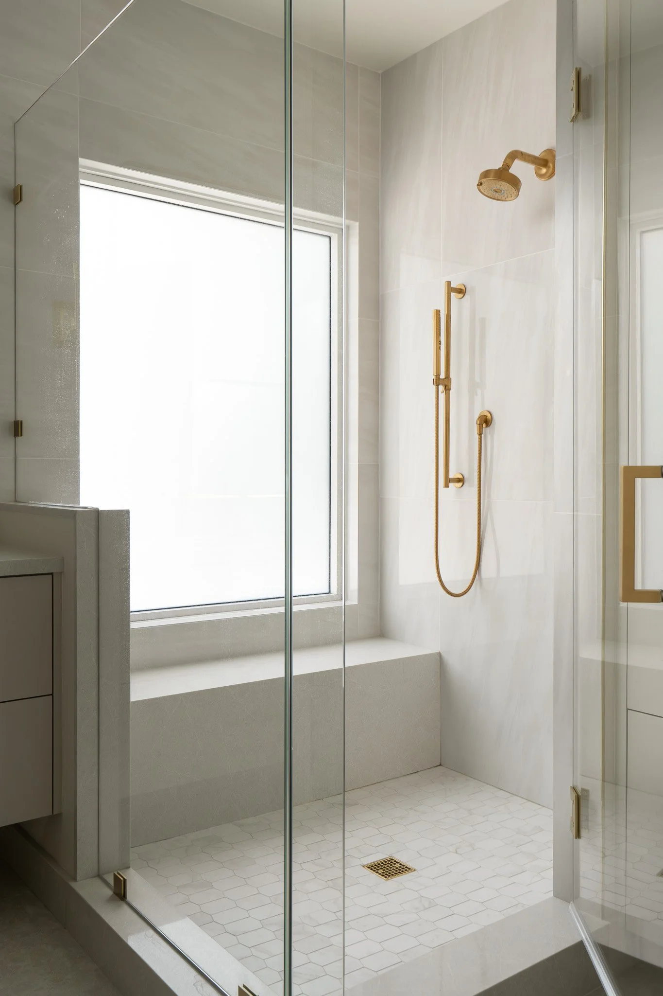 A modern shower area with a glass enclosure, a gold showerhead, and a handheld shower fixture on a marble wall, with a large window providing natural light.