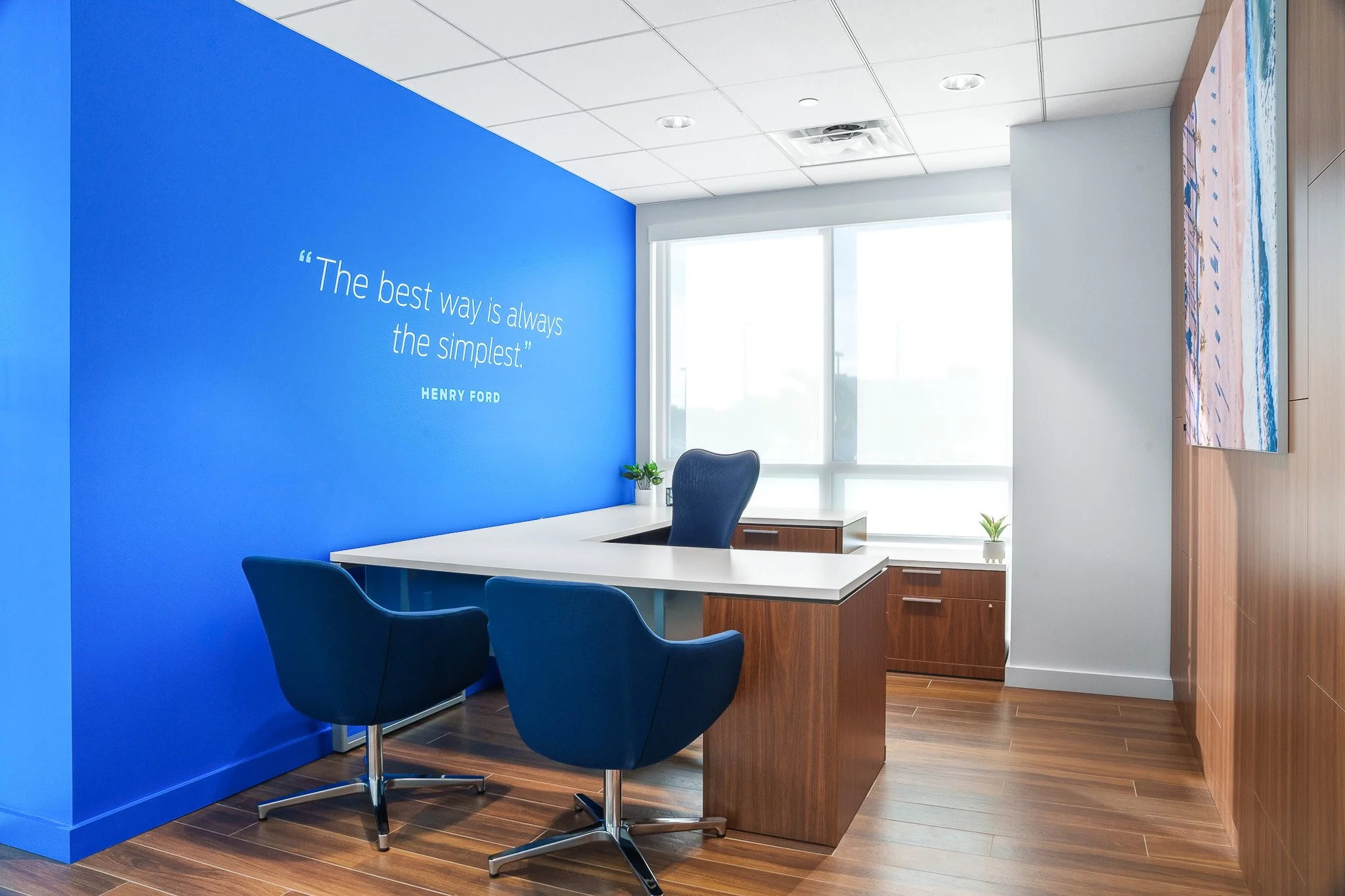 An office with a blue accent wall, quote by Henry Ford, a white desk, black office chairs, large windows, some plants, and abstract art on the wall.