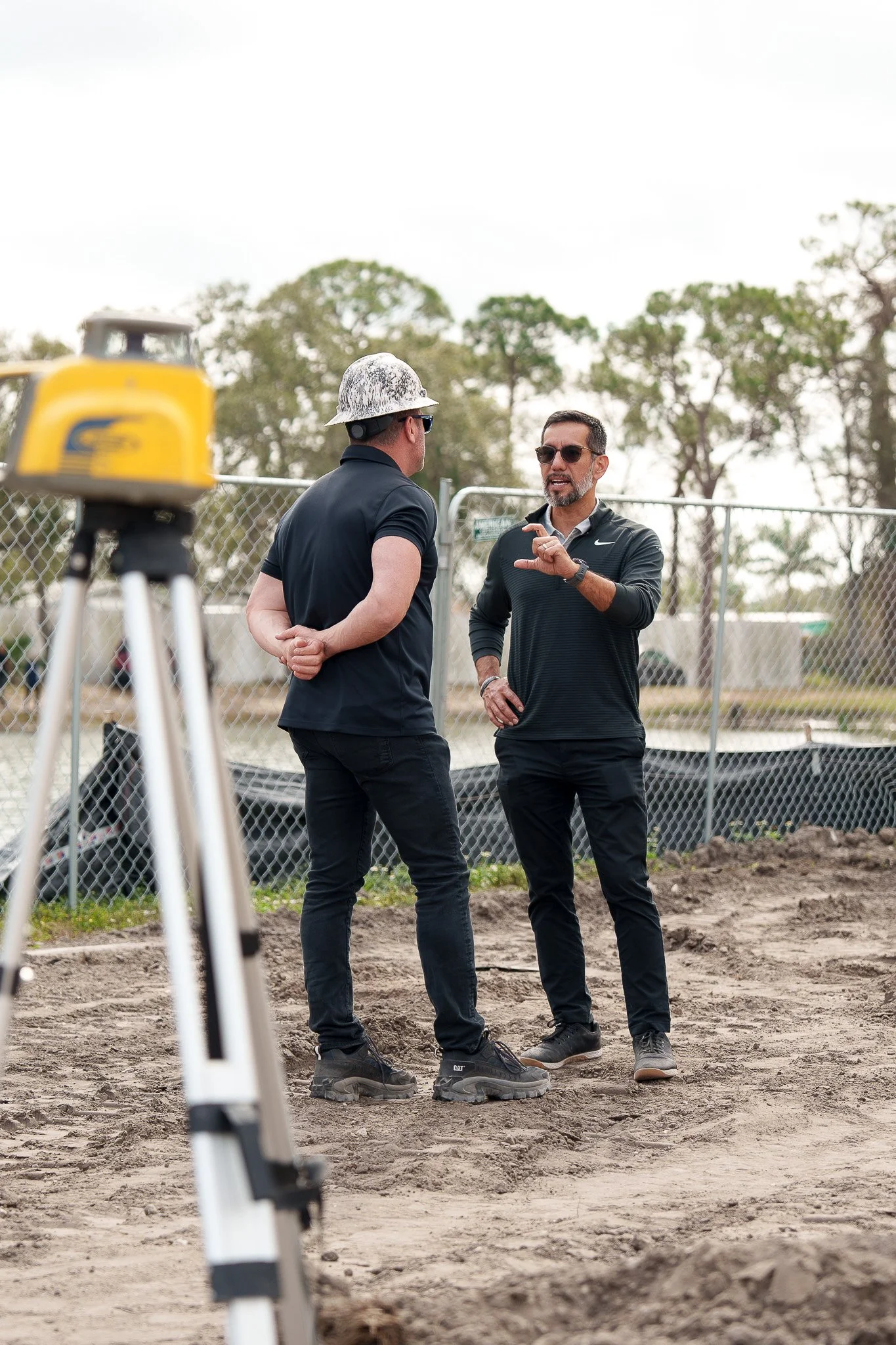 Two men in black clothing and sunglasses having a conversation outdoors, with a construction site or fenced area and trees in the background. One wears a construction helmet and the other a wristwatch. There is surveying equipment in the foreground.