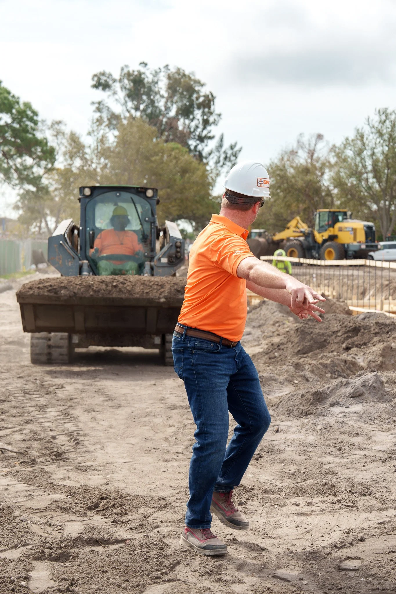 A construction site with a man in an orange shirt and blue jeans guiding a man in a yellow hard hat and orange safety vest operating a skid steer loader, with trees in the background and cloudy sky overhead.