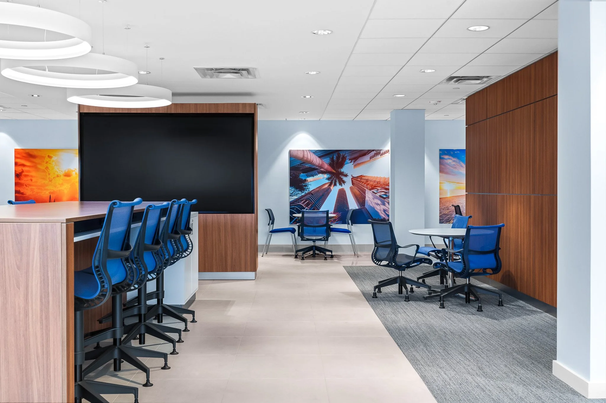 Modern office meeting room with blue chairs, wall art of cityscapes, a large blackboard or screen, and white ceiling with circular light fixtures.