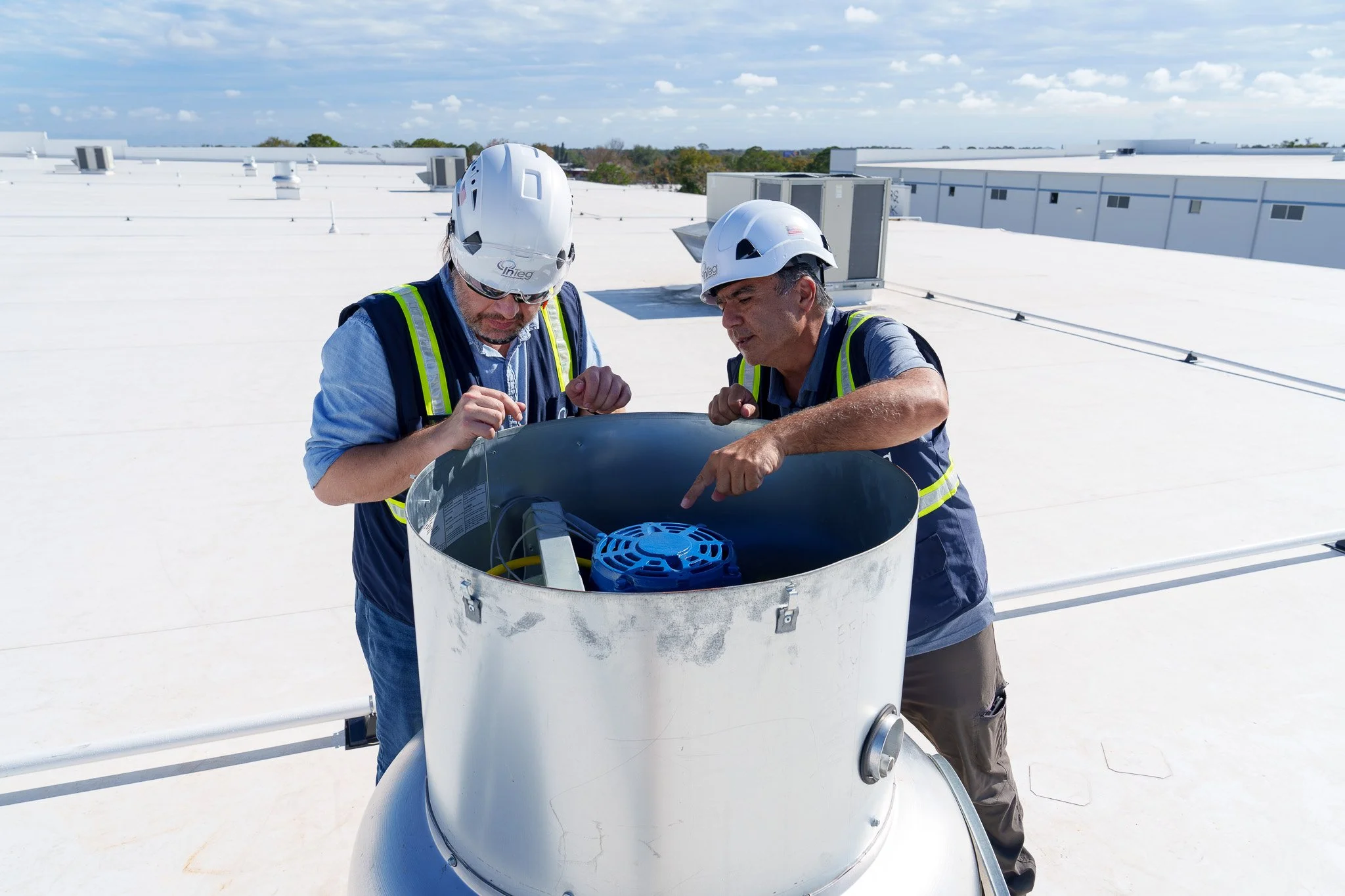 Two workers wearing safety helmets and vests inspecting equipment on a white rooftop under a partly cloudy sky.