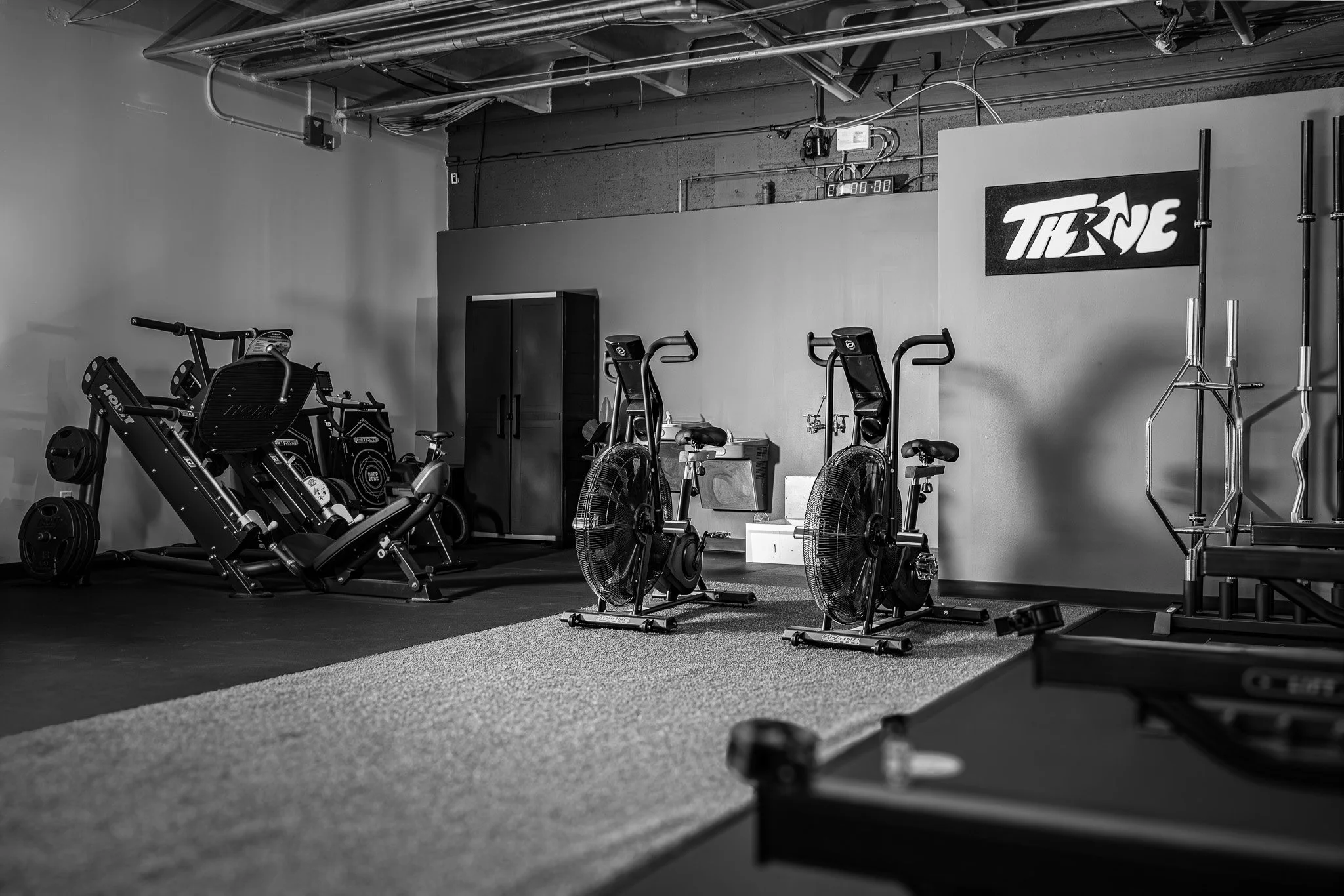 Fitness gym with exercise equipment including rowing machines, stationary bikes, and a rack of weights against concrete walls, with a "THRIVE" sign on the wall.