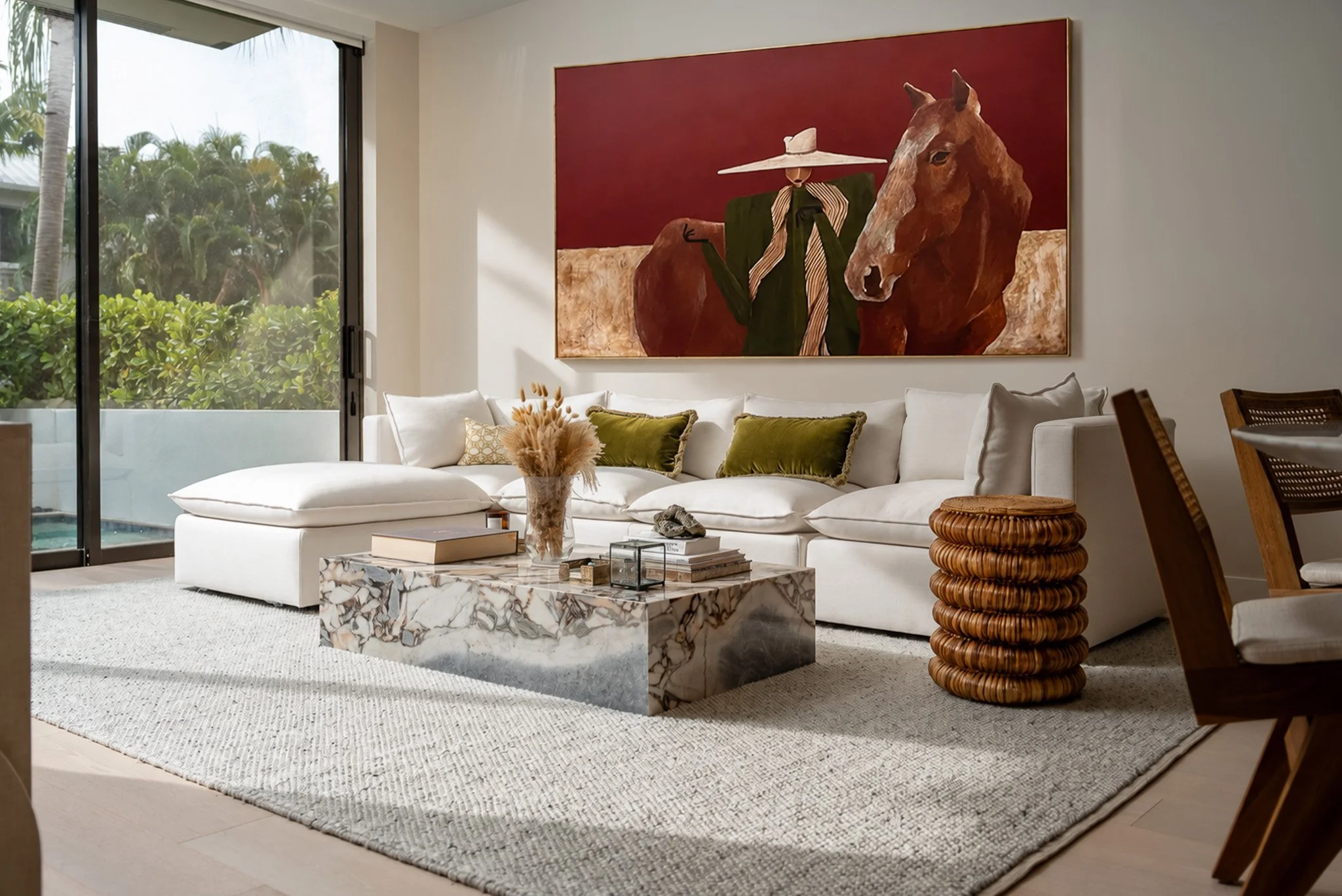 Modern living room with white sectional sofa, green pillows, abstract marble coffee table, large painting of a person with a horse, tropical plants outside sliding glass door.