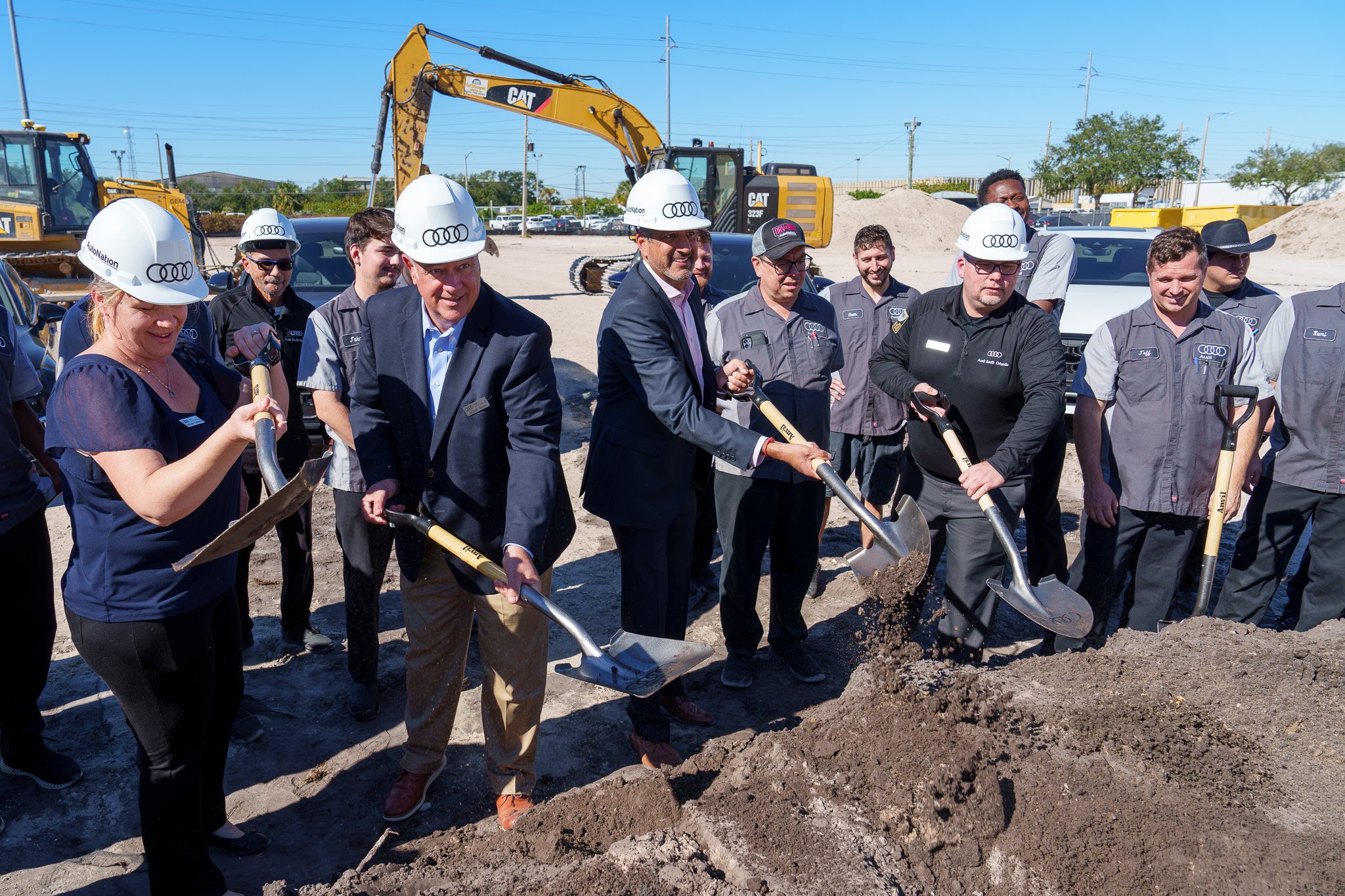 Group of people at a groundbreaking ceremony holding shovels in a construction site. Some are wearing hard hats with the Audi logo, and there are construction equipment and vehicles in the background.
